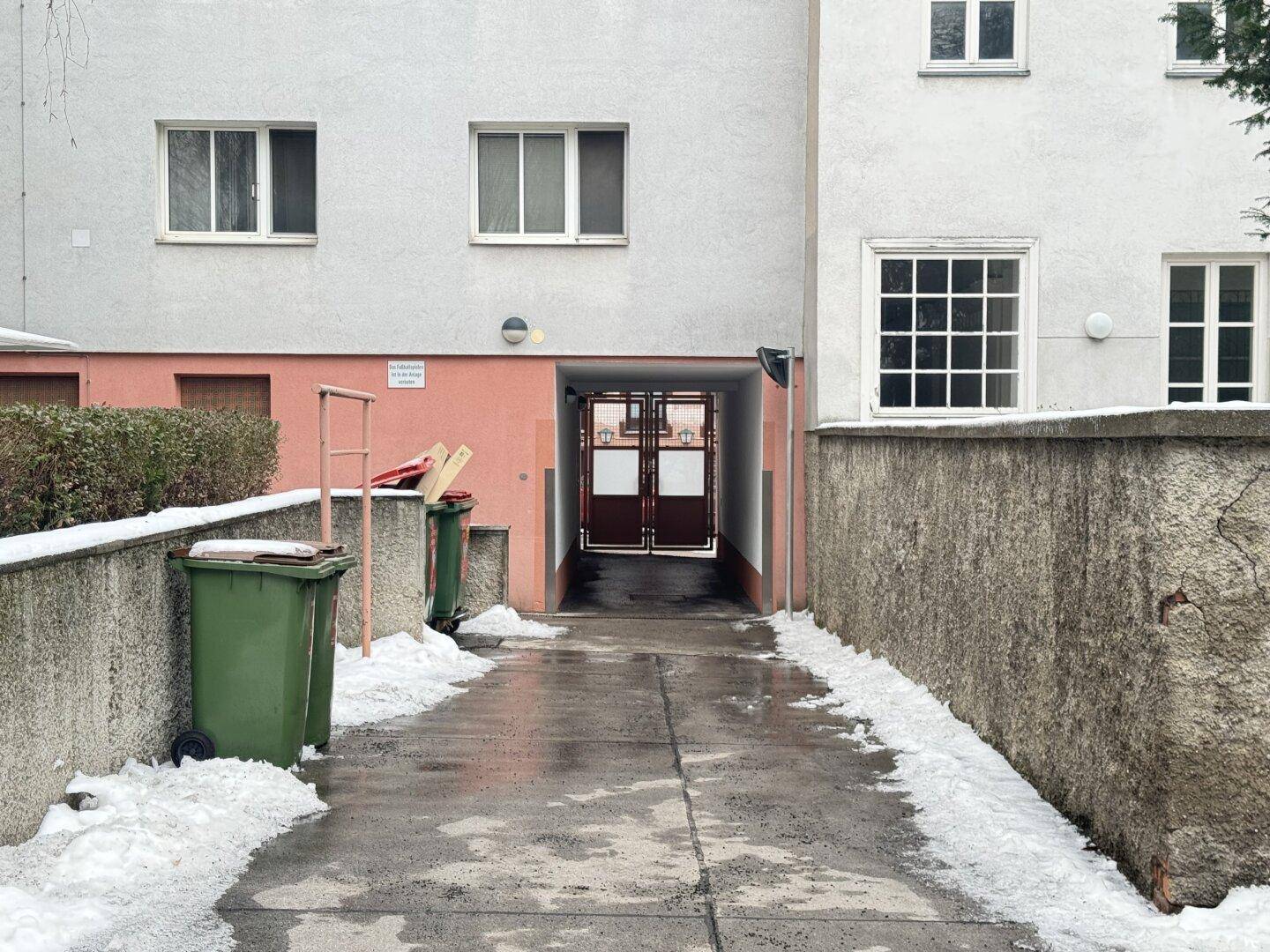 Gebäudeeingang mit Durchgang, umgeben von Mauern, Schnee und Mülltonnen im Außenbereich.