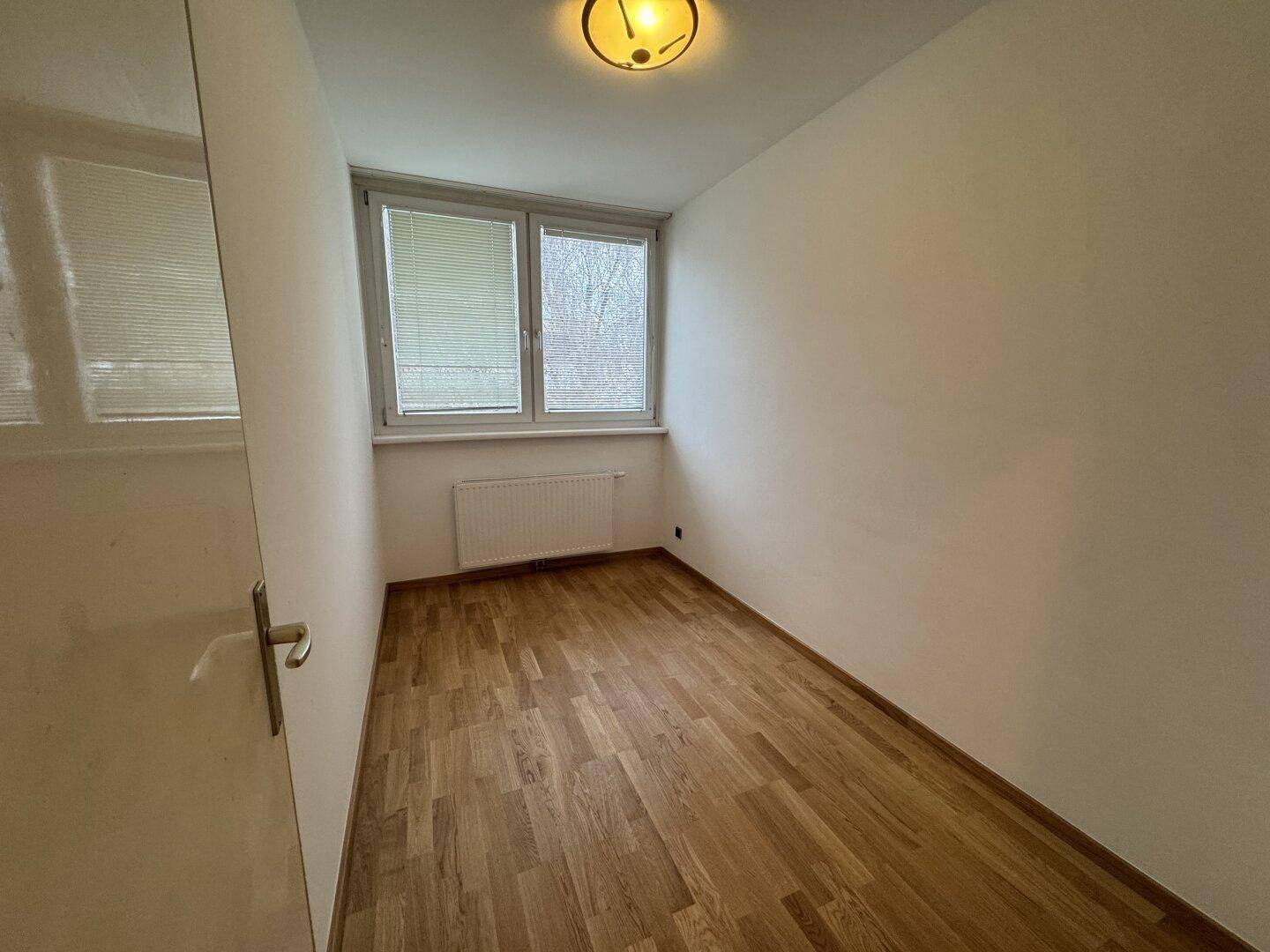 Helles, leeres Zimmer mit Parkettboden und Fensterblick ins Grüne, ideal als Schlaf- oder Arbeitszimmer.