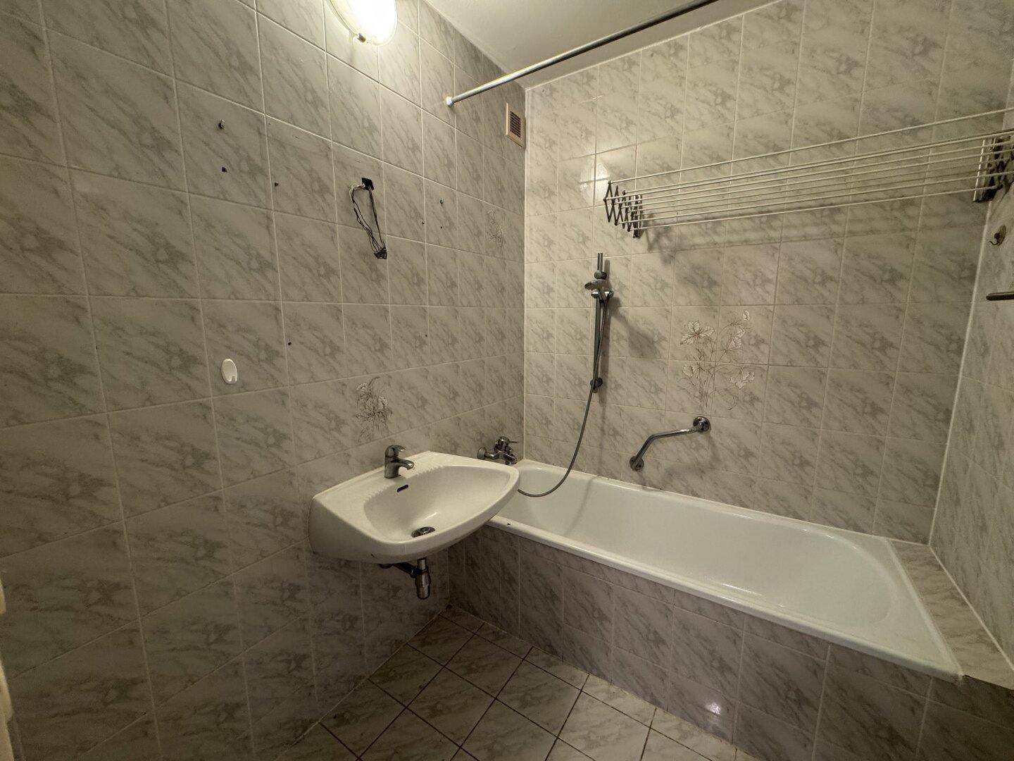 Älteres Badezimmer mit Badewanne, Waschbecken und hellen Fliesen, das eine Modernisierung benötigt.