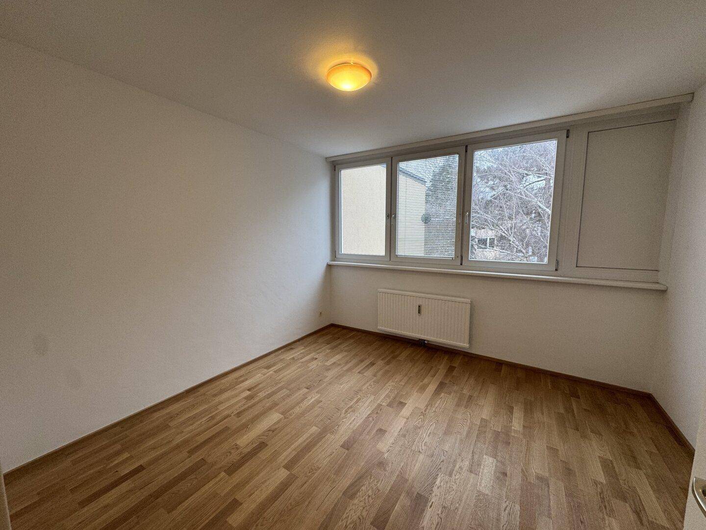 Geräumiges, leeres Zimmer mit Parkettboden und großen Fenstern, bietet viel Tageslicht und Ausblick.