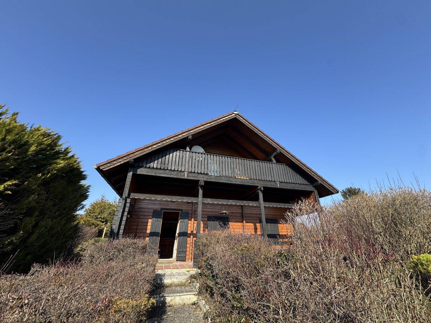 Charmantes Holzhaus mit Balkon und Satteldach, umgeben von grüner Vegetation und Büschen.