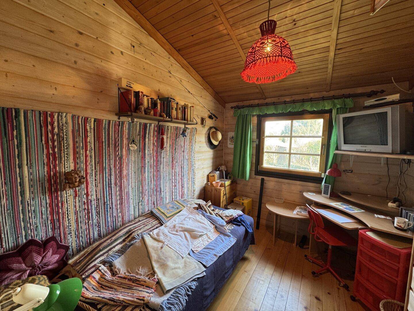 Kleines Schlafzimmer im Dachgeschoss mit Holzwänden, Schreibtisch und Fenster mit Blick ins Grüne.