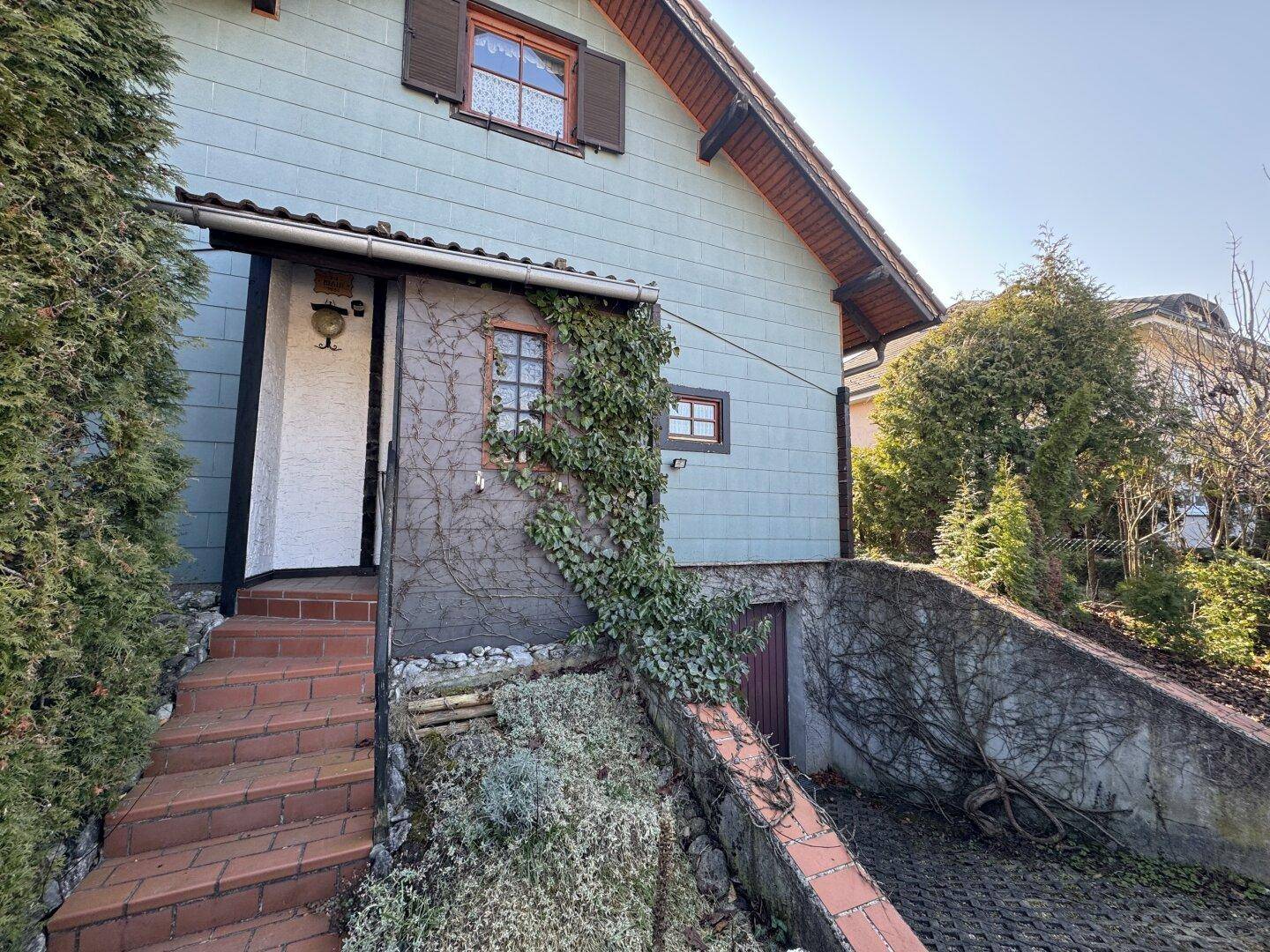 Eingangsbereich eines Hauses mit Steintreppe und bewachsenen Fassadenbereichen, umgeben von Grün.