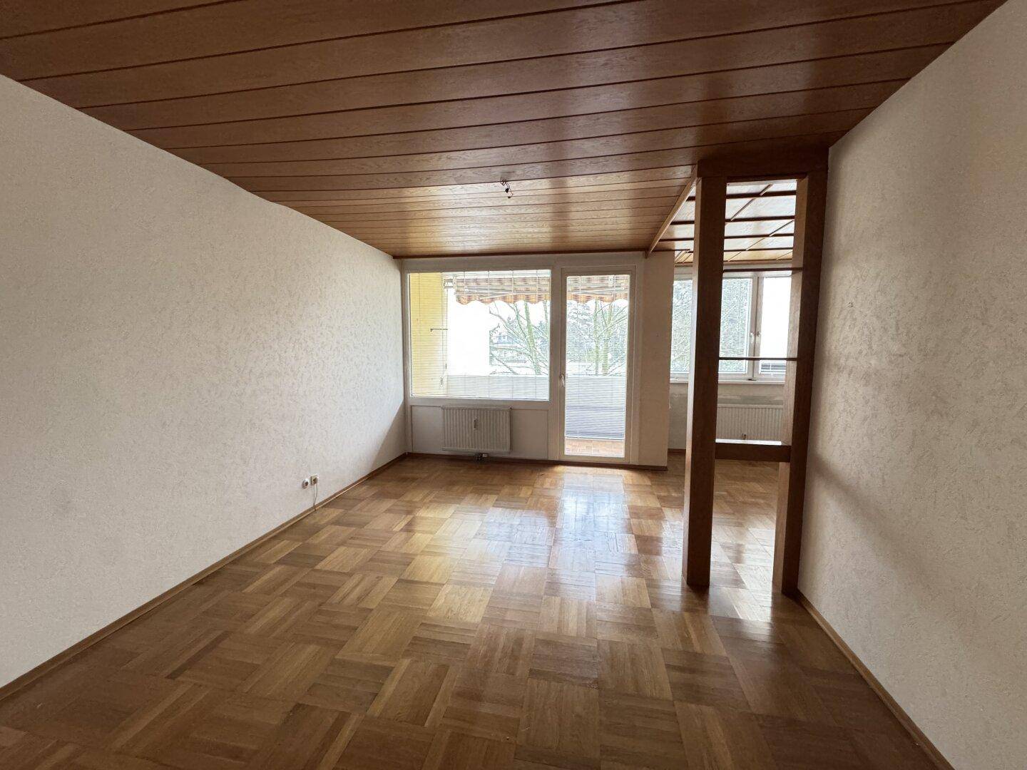 Geräumiges Wohnzimmer mit Parkettboden, Holzdecke und Zugang zu einem Balkon.