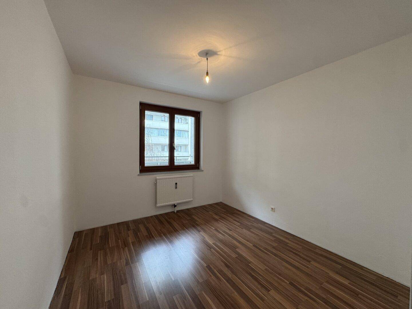 Schlafzimmer mit Holzboden, weißen Wänden und einem Fenster, das Tageslicht hereinlässt.