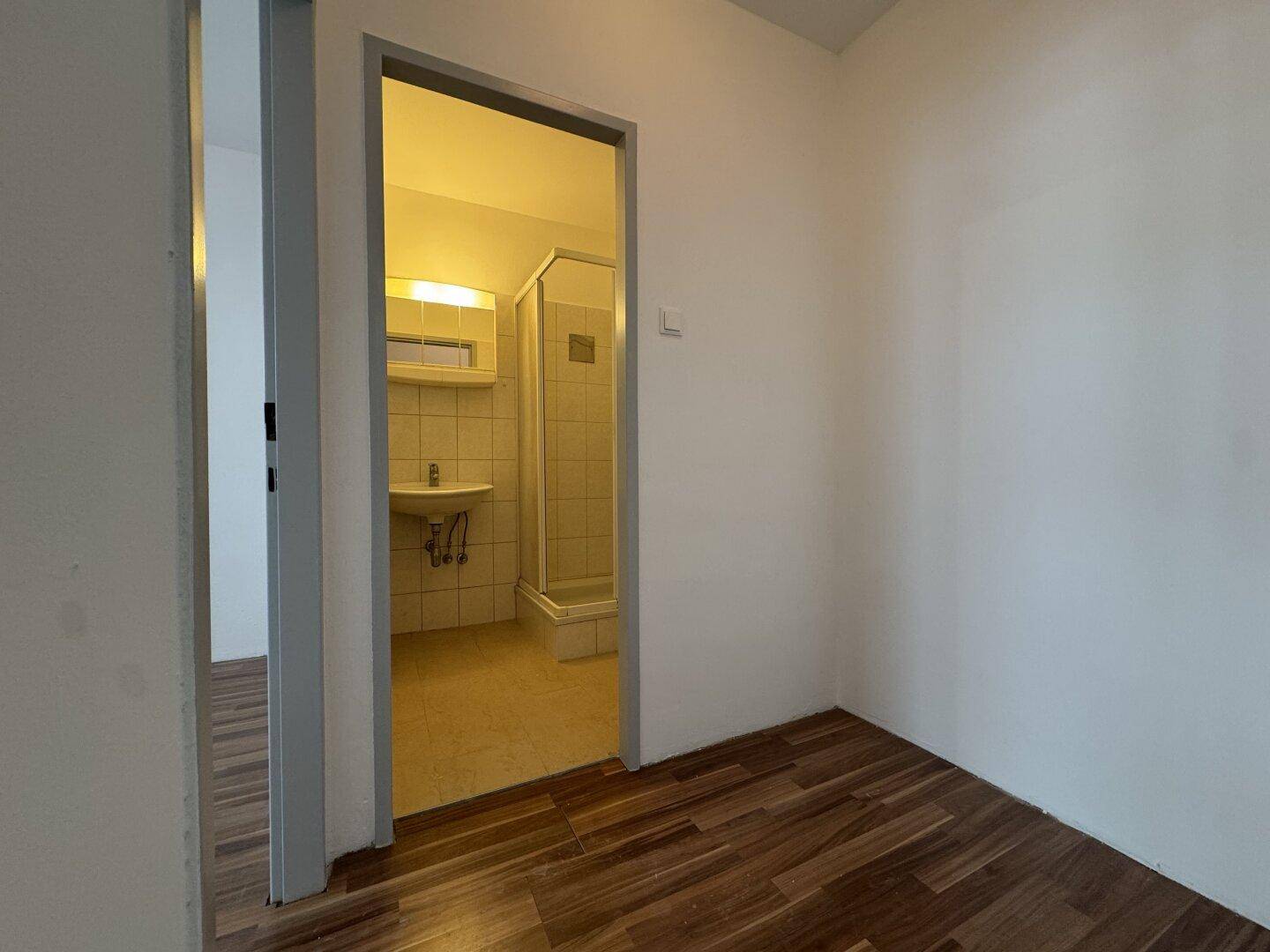 Blick von einem Zimmer mit Holzboden in ein Badezimmer mit Duschkabine und Waschbecken.