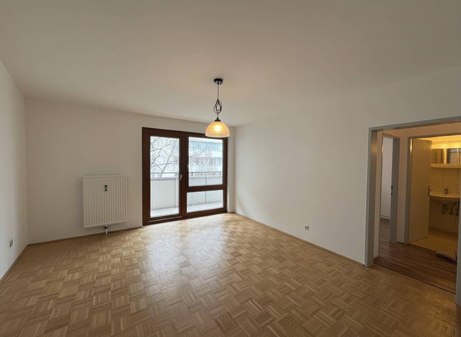 Helles Wohnzimmer mit Parkettboden und großem Fenster, das Zugang zu einem Balkon bietet.