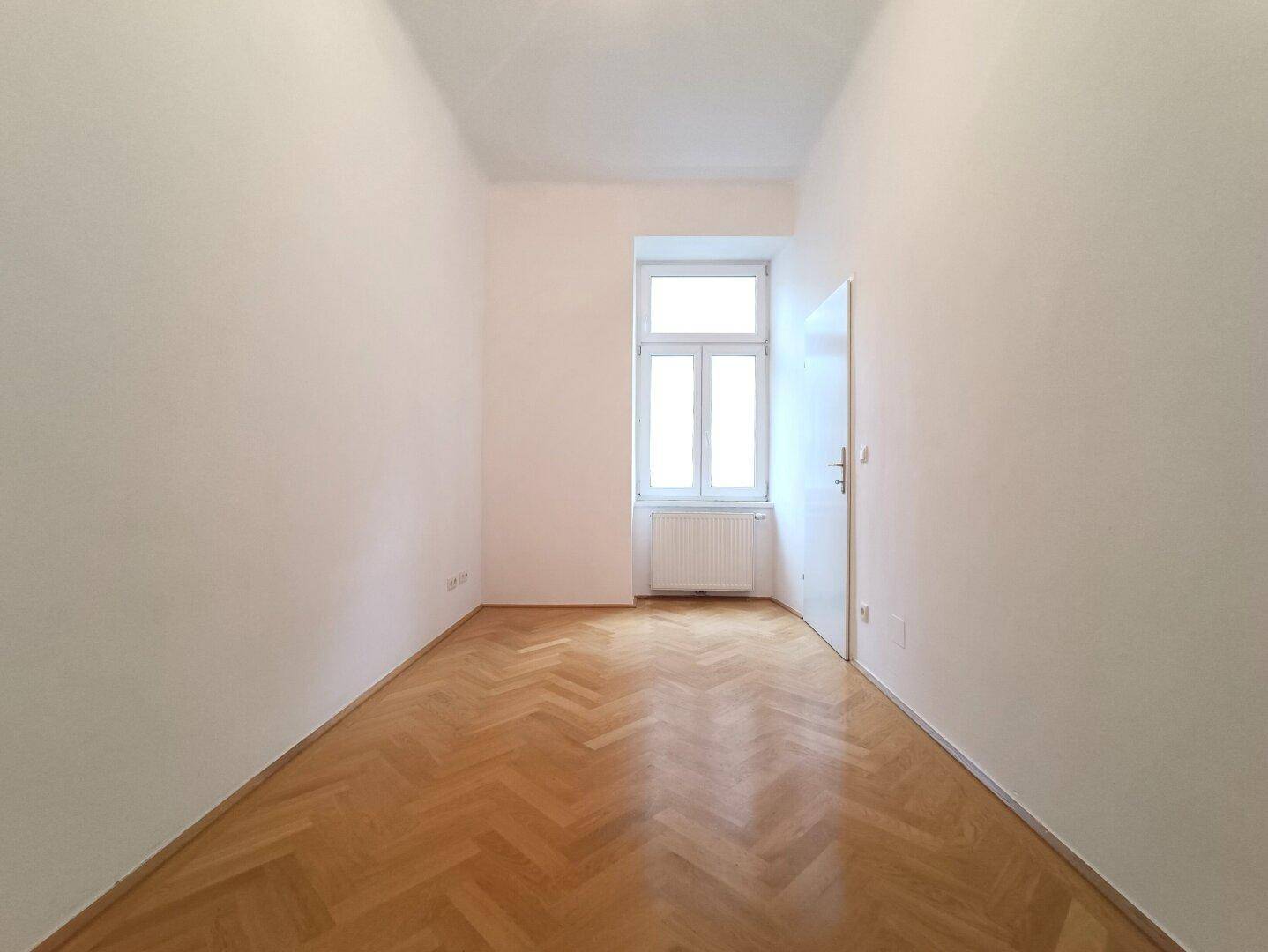 Schmales Zimmer mit Fischgrätparkett, einem Fenster und weiß gestrichenen Wänden.