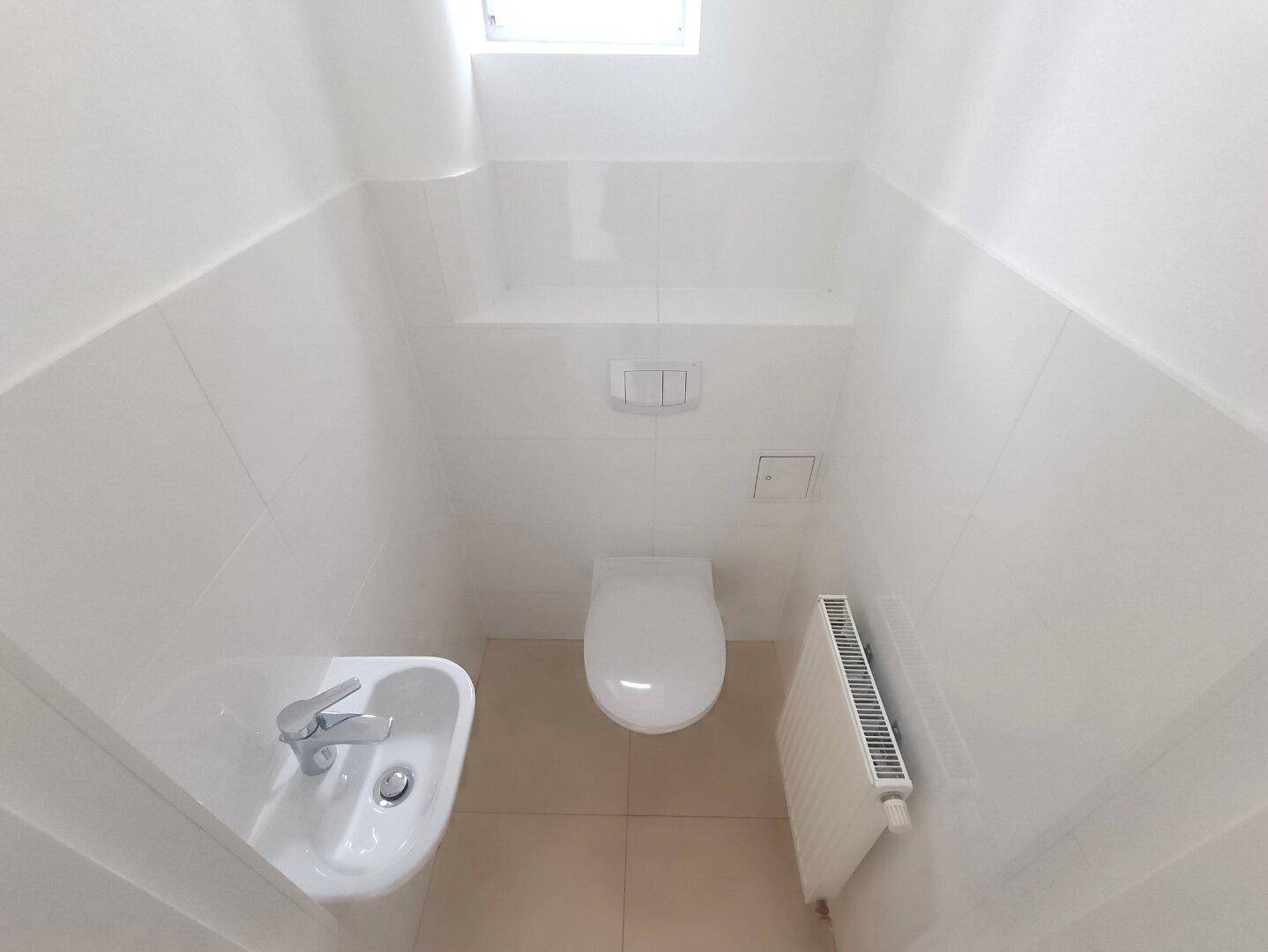 Separates WC mit wandhängender Toilette, kleinem Waschbecken und einem Fenster für Tageslicht.