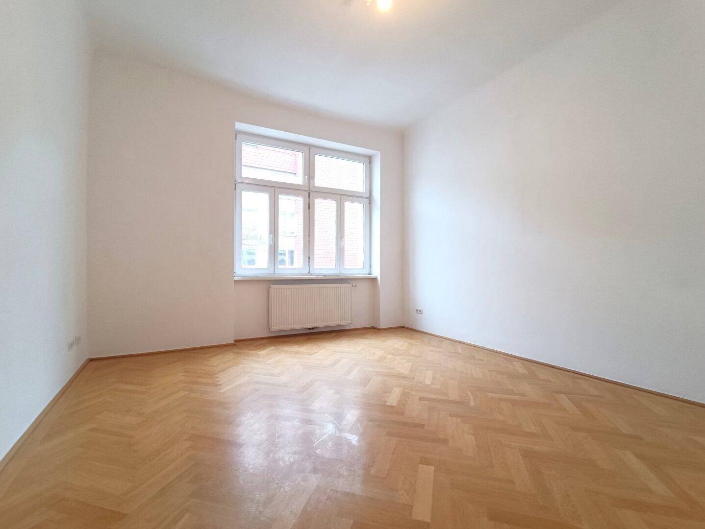 Leeres Zimmer mit hellem Fischgrätparkettboden und großem Fenster für viel Tageslicht.