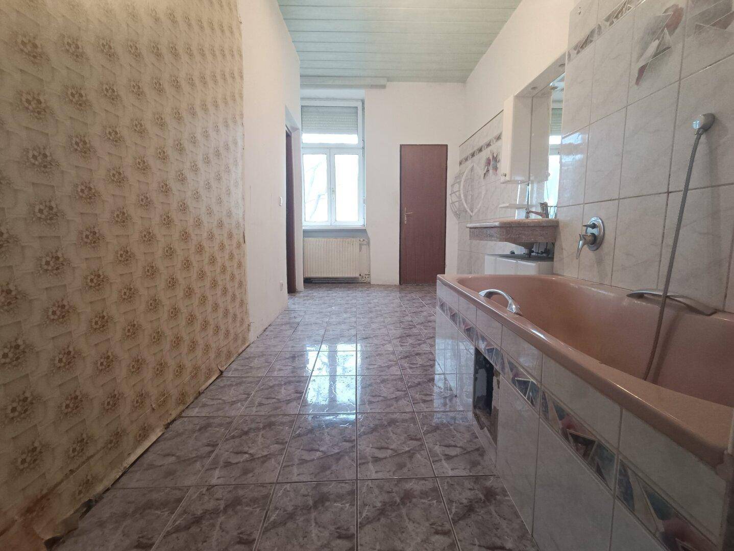 Geräumiges Badezimmer mit Badewanne, Fliesenboden und Fenster, renovierungsbedürftig.