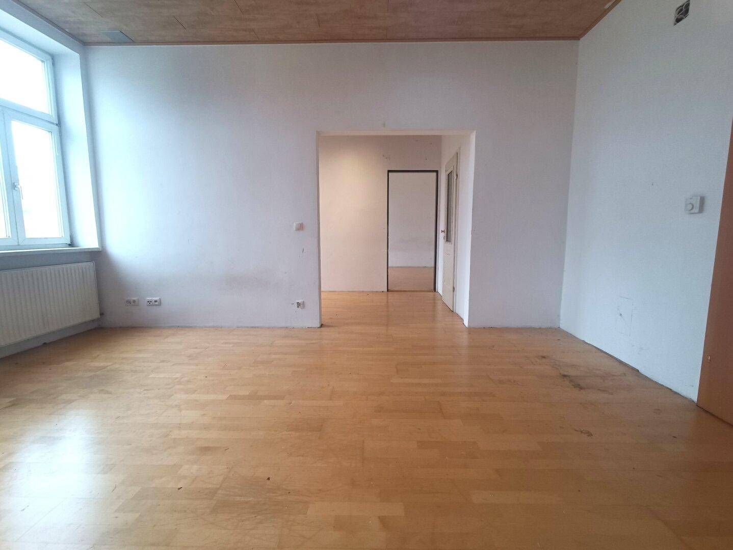 Heller Raum mit großem Fenster, Parkettboden und Zugang zu weiteren Zimmern.
