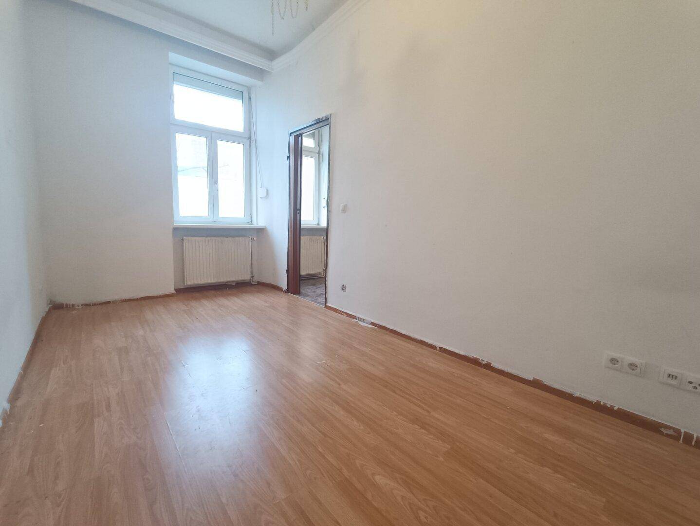 Helles Zimmer mit Laminatboden, großem Fenster und Renovierungsbedarf.
