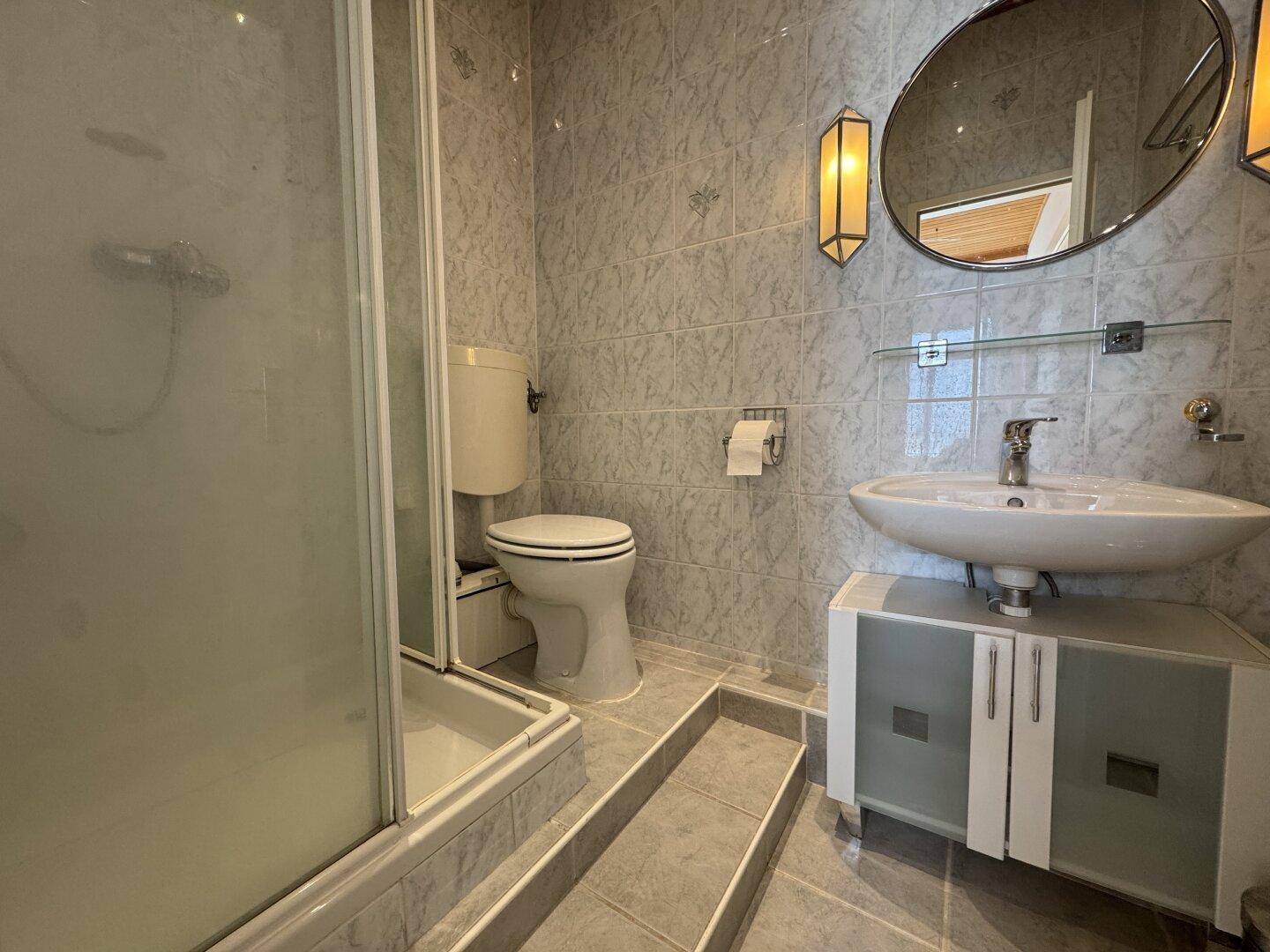 Gefliestes Badezimmer mit Dusche, WC und Waschbecken in älterem Zustand.