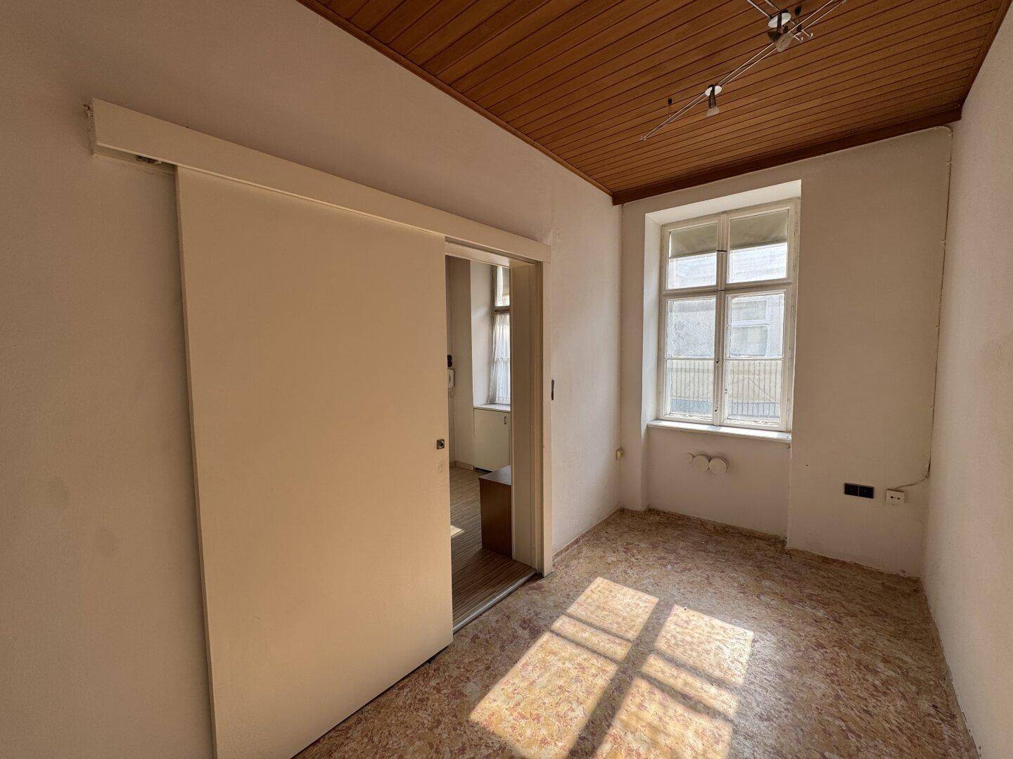 Helles Zimmer mit Holzdecke, Fenster und Schiebetür, sanierungsbedürftig.