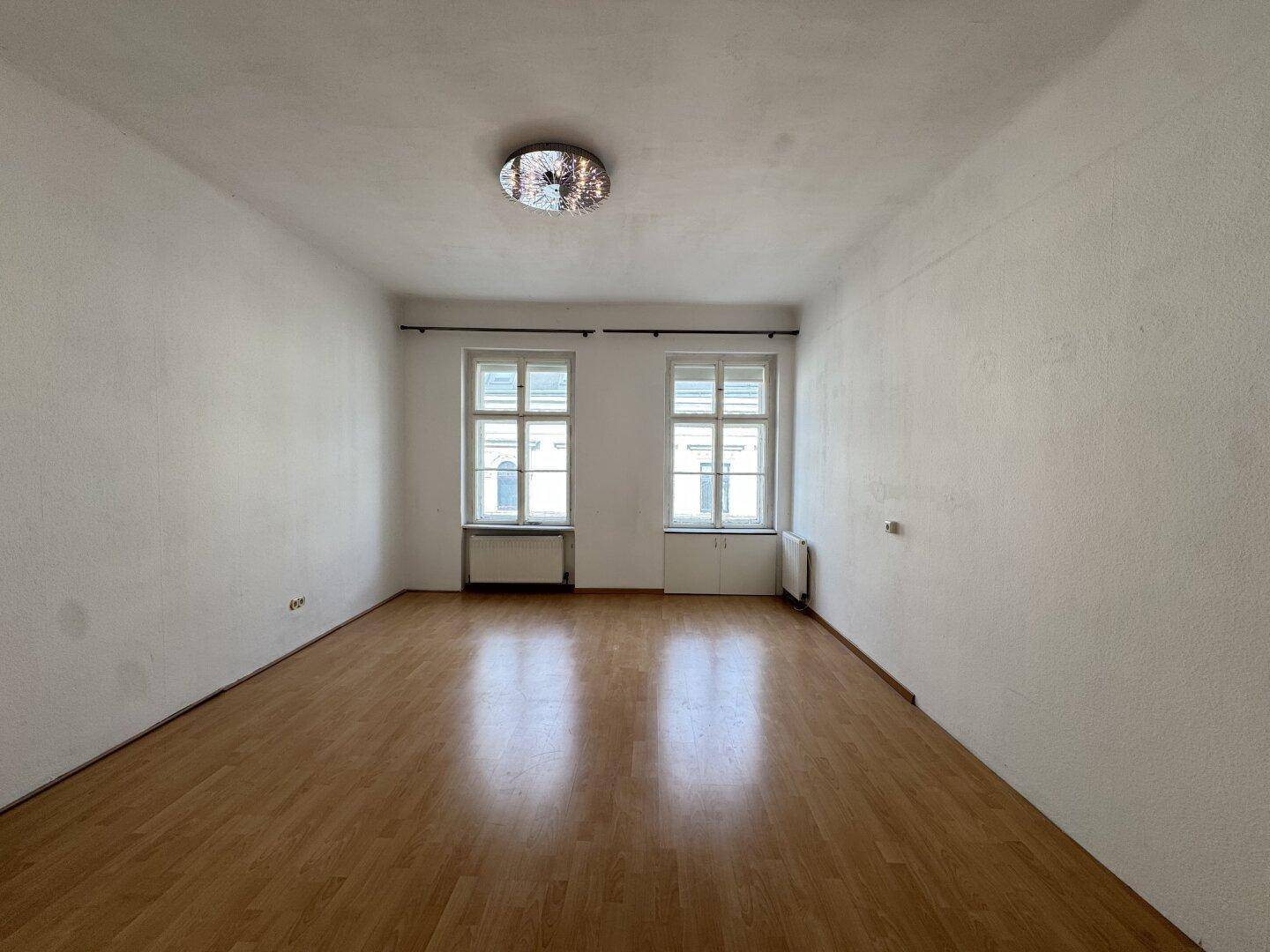 Helles leeres Zimmer mit Parkettboden und zwei großen Fenstern.