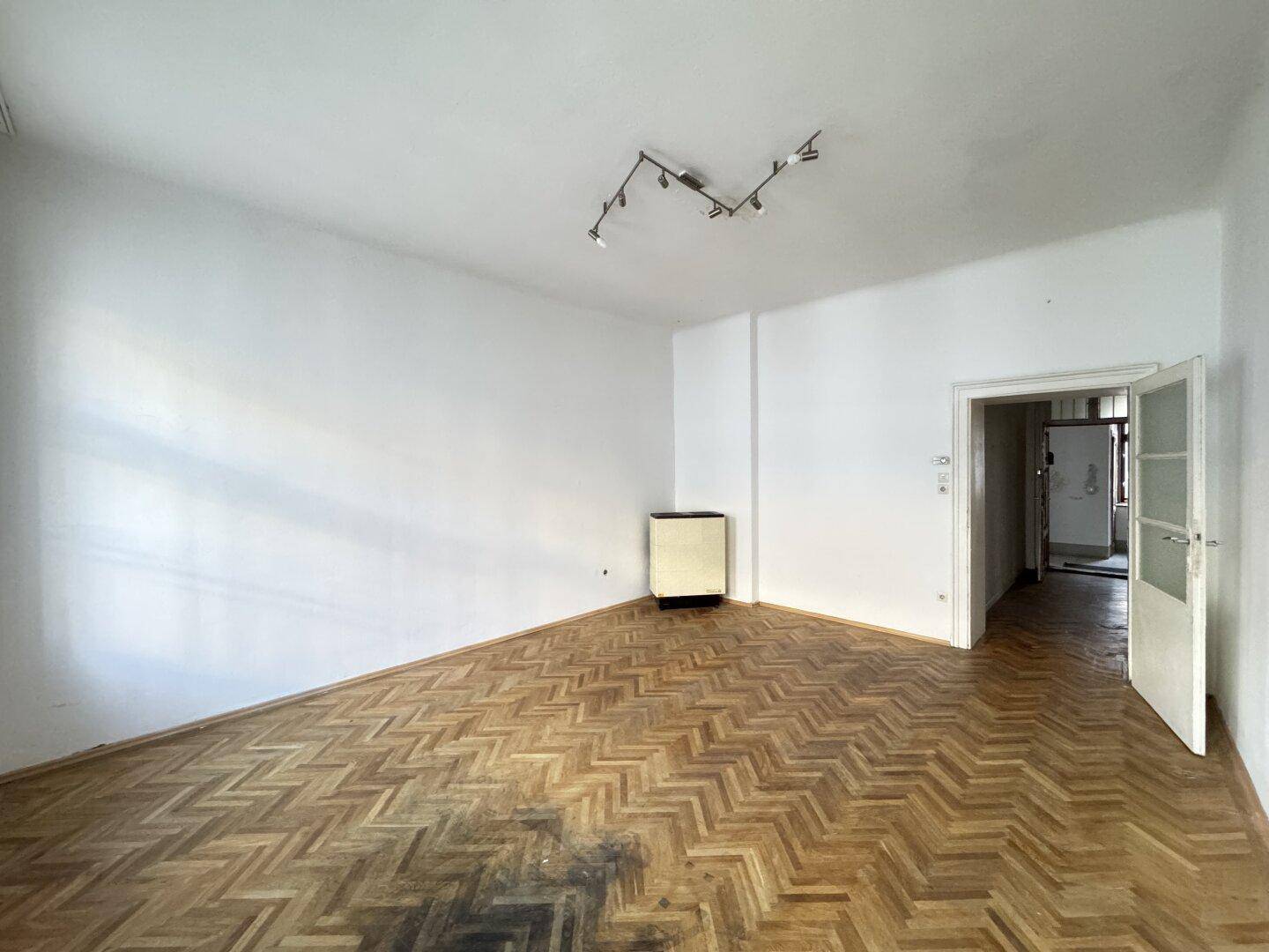 Leeres Zimmer mit Fischgrätparkett und Heizkörper, sanierungsbedürftig.