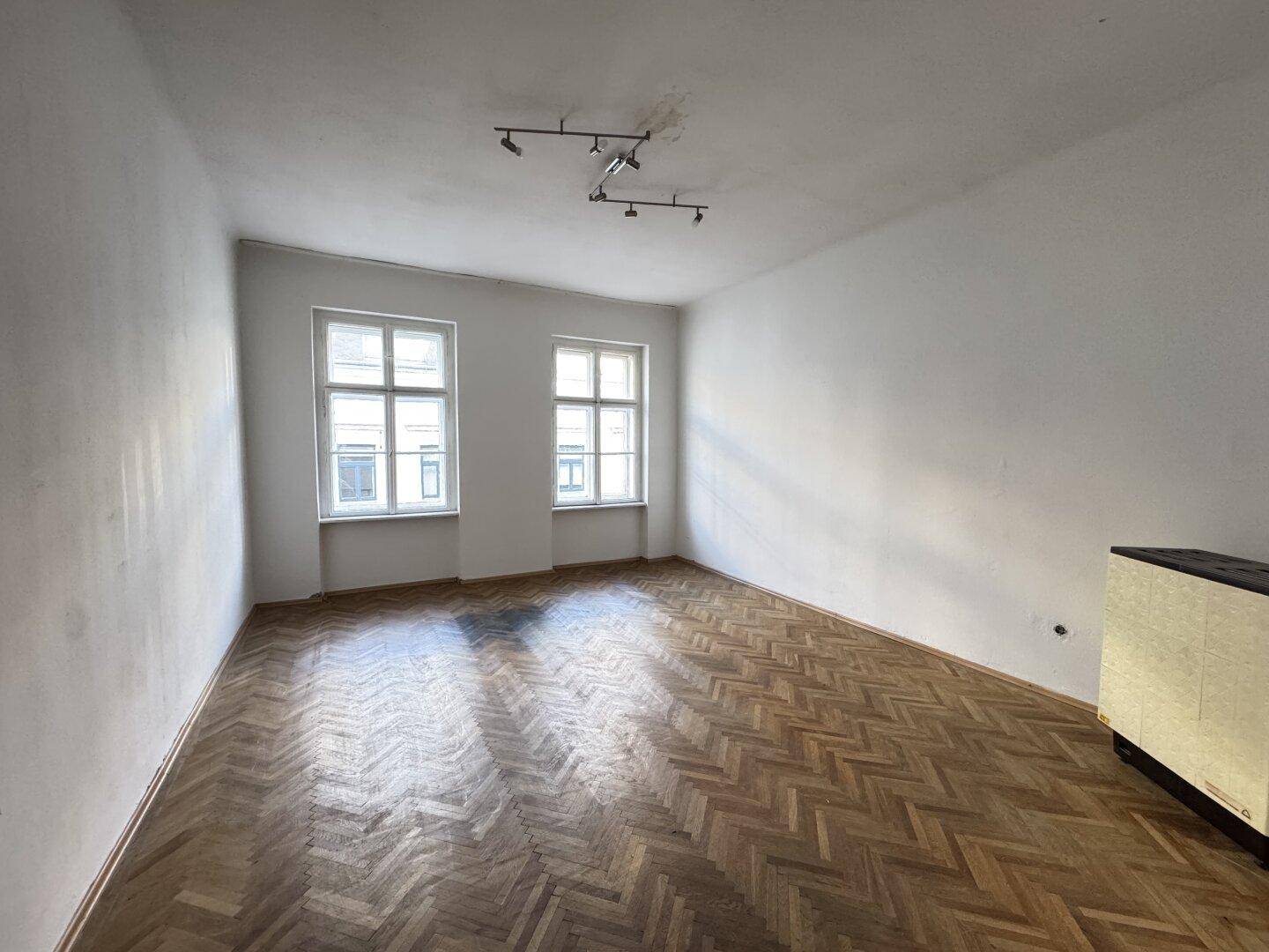 Helles leeres Zimmer mit Fischgrätparkett und zwei großen Fenstern.