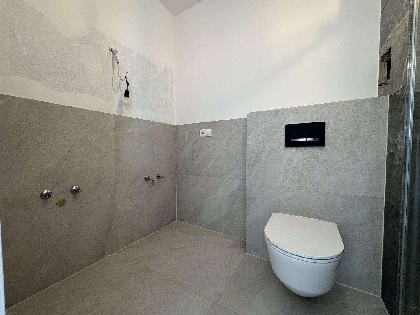 Badezimmer im Bau mit Wand-WC, grauen Fliesen und Vorbereitung für den Waschtisch.