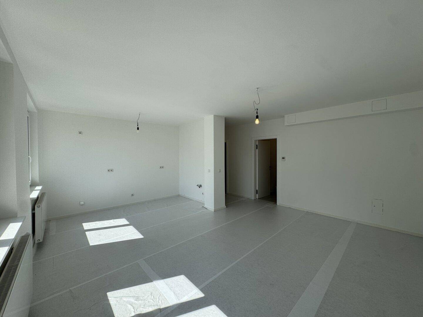 Großer, heller Raum mit viel Tageslicht, abgedecktem Boden und Zugang zu weiteren Zimmern.