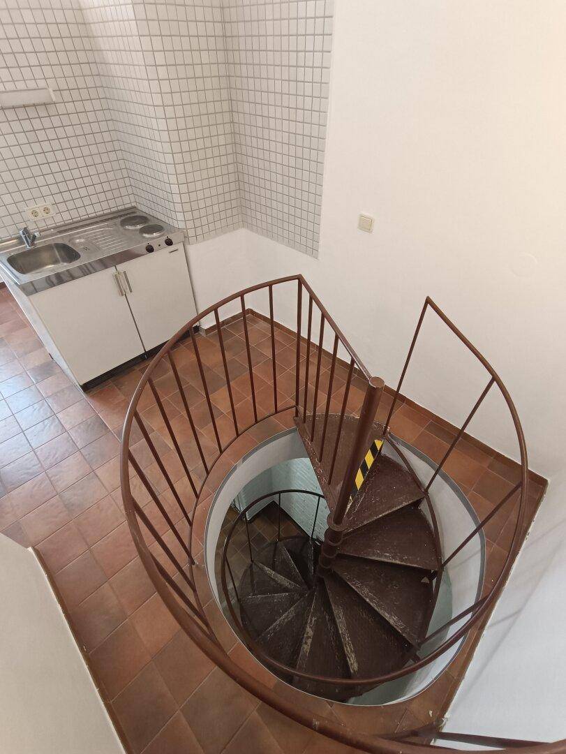 Blick von oben auf eine Wendeltreppe in einem Raum mit Küchenzeile und Terrakottafliesen.