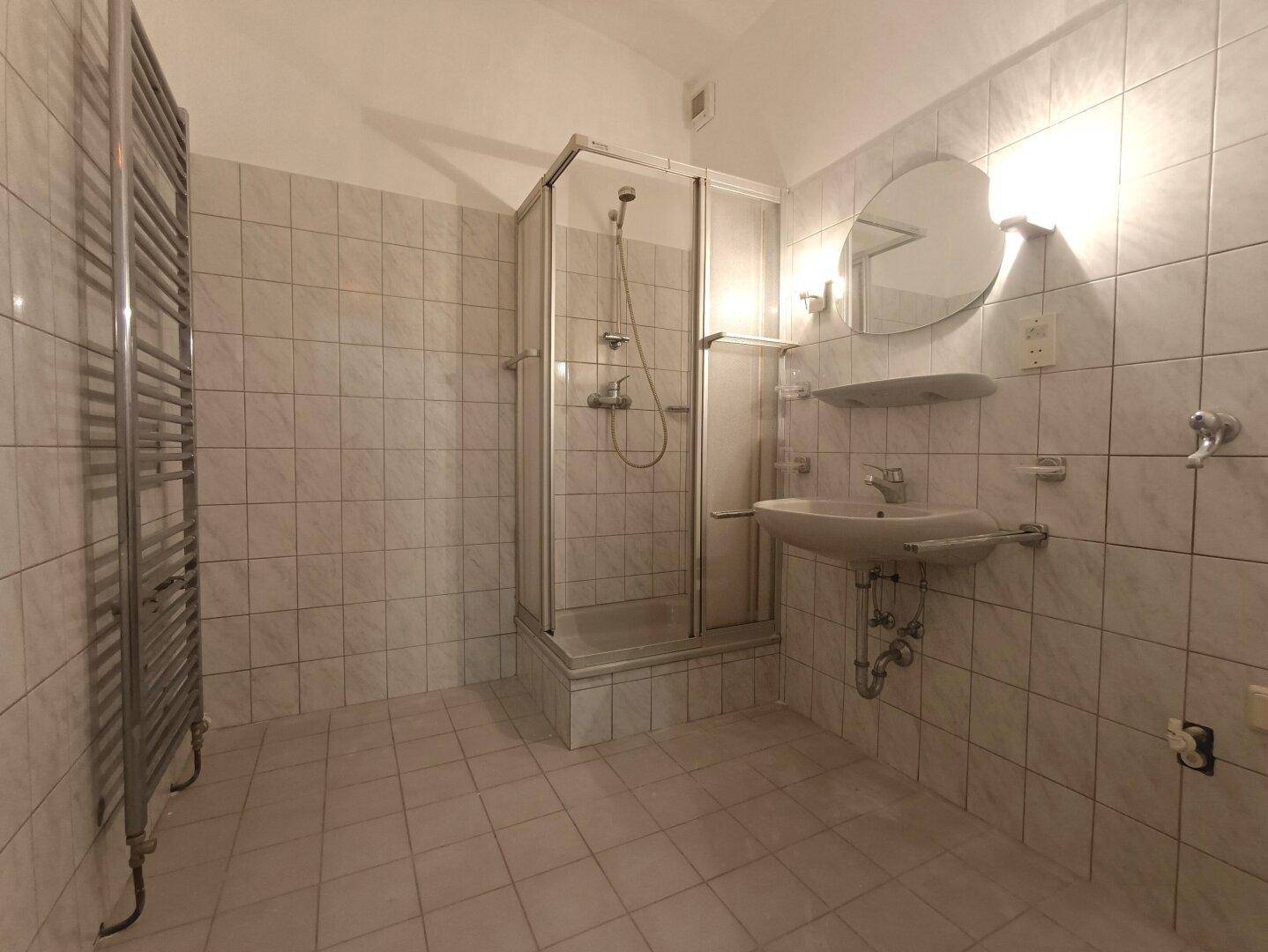 Badezimmer mit weißen Fliesen, einer Eckduschkabine, Waschbecken und Handtuchheizkörper.