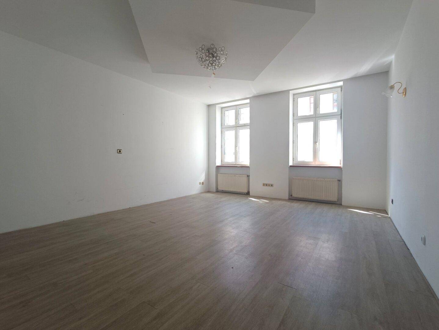 Geräumiges Wohnzimmer mit Laminatboden, zwei großen Fenstern und hohen Decken.