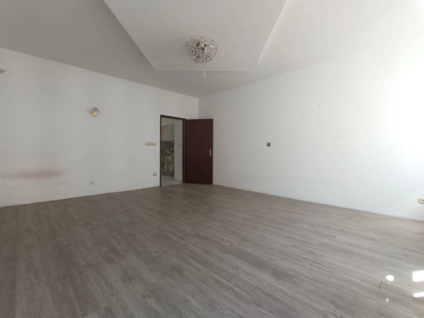 Heller Wohnbereich mit Laminatboden, hohen Decken und moderner Deckenbeleuchtung.