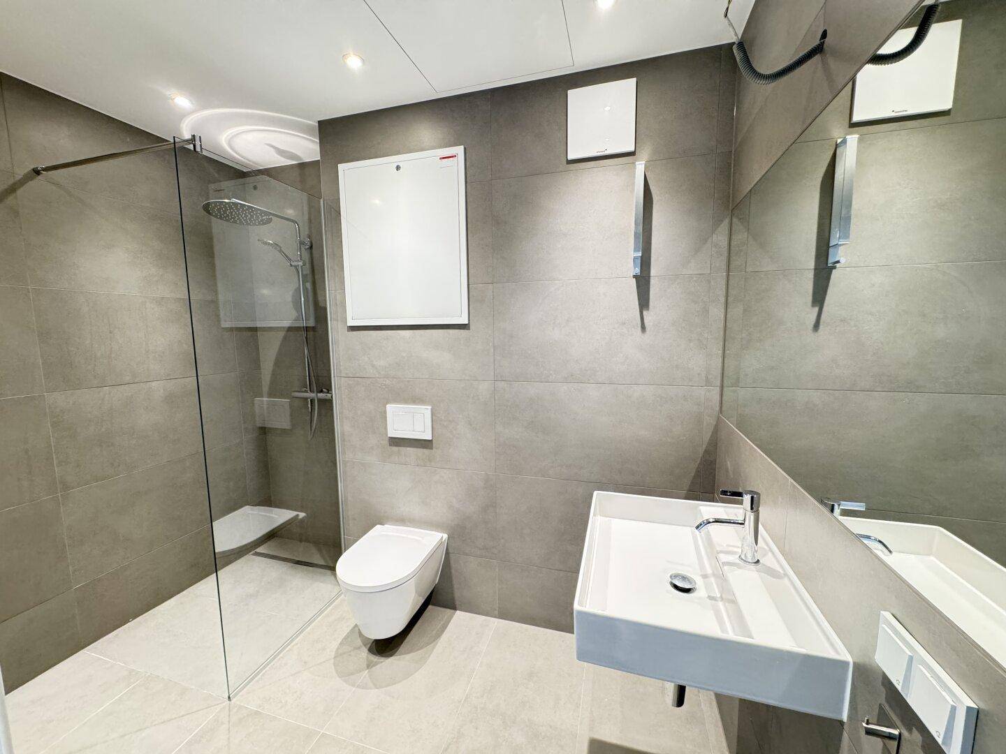 Modernes Badezimmer mit grauen Fliesen, bodengleicher Dusche und wandhängendem WC.