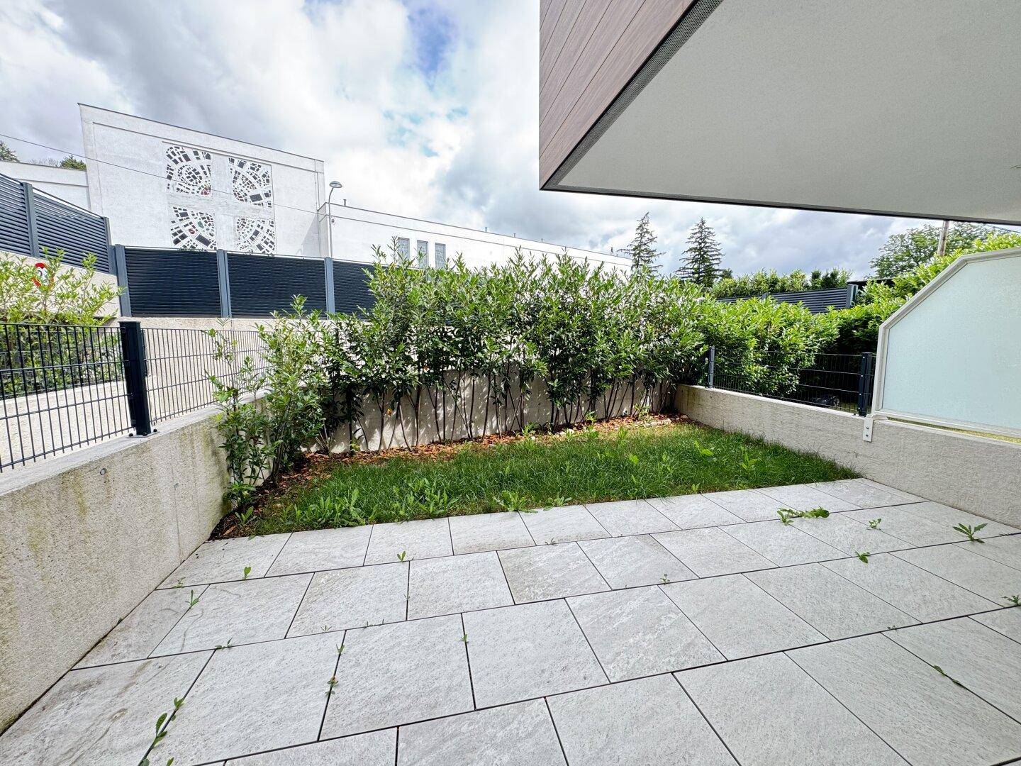 Kleine Terrasse mit angrenzender Rasenfläche und Hecke, umgeben von modernen Gebäuden.