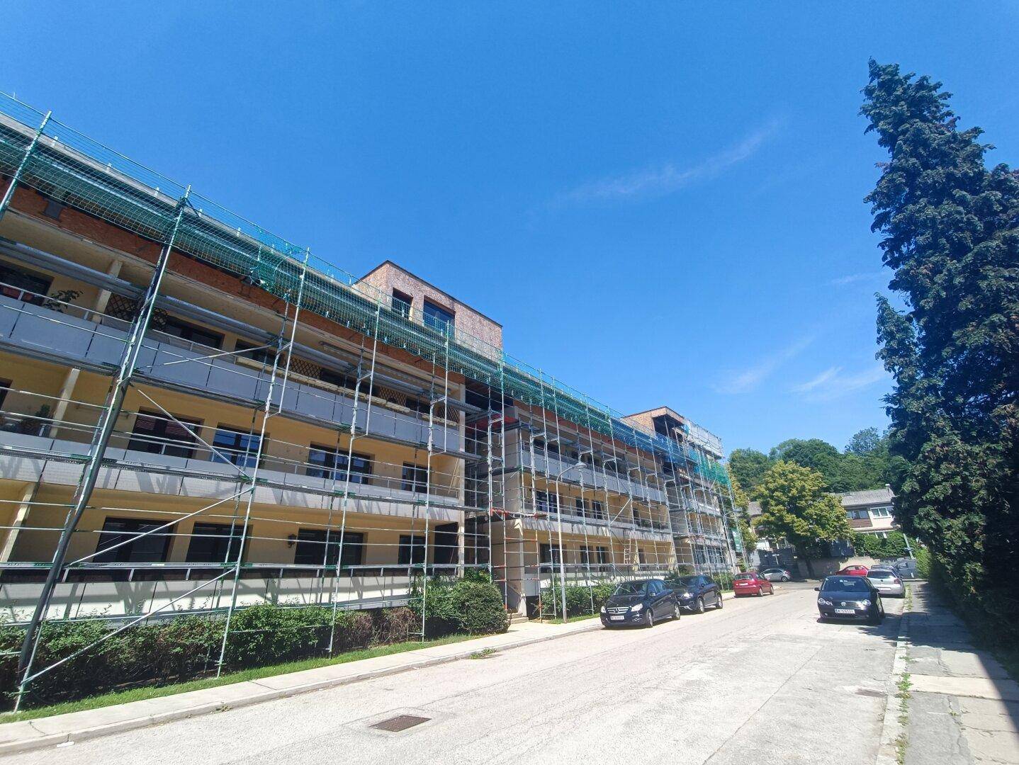 Mehrfamilienhaus mit Gerüst an der Fassade und parkenden Autos auf der Straße.