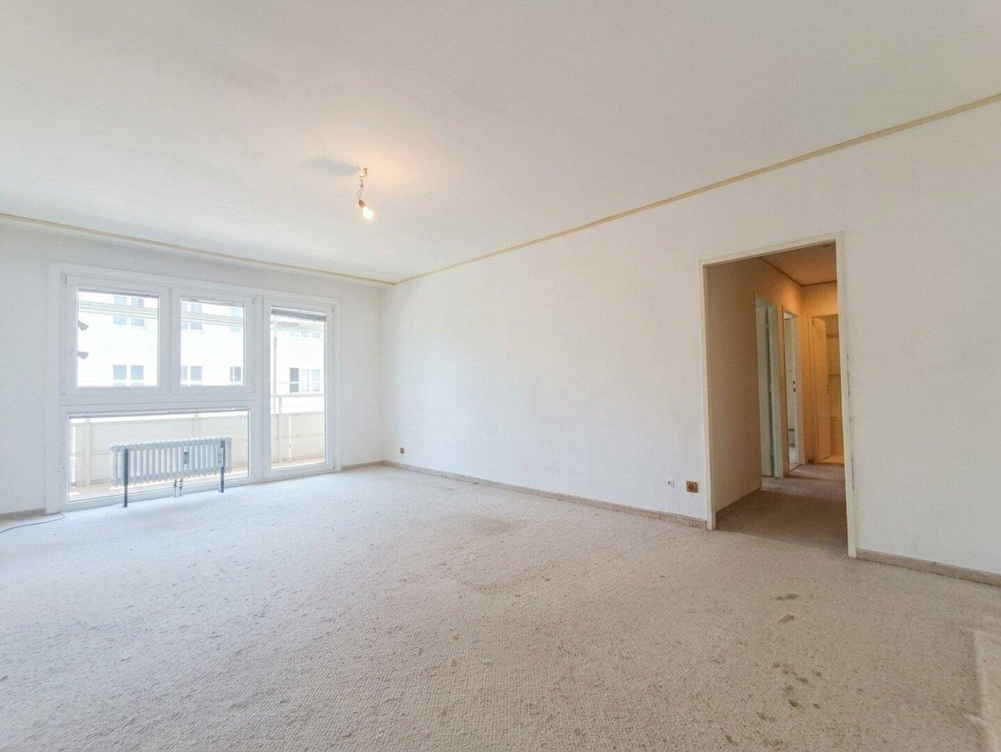 Leeres Wohnzimmer mit Teppichboden und Zugang zu einem kleinen Balkon.