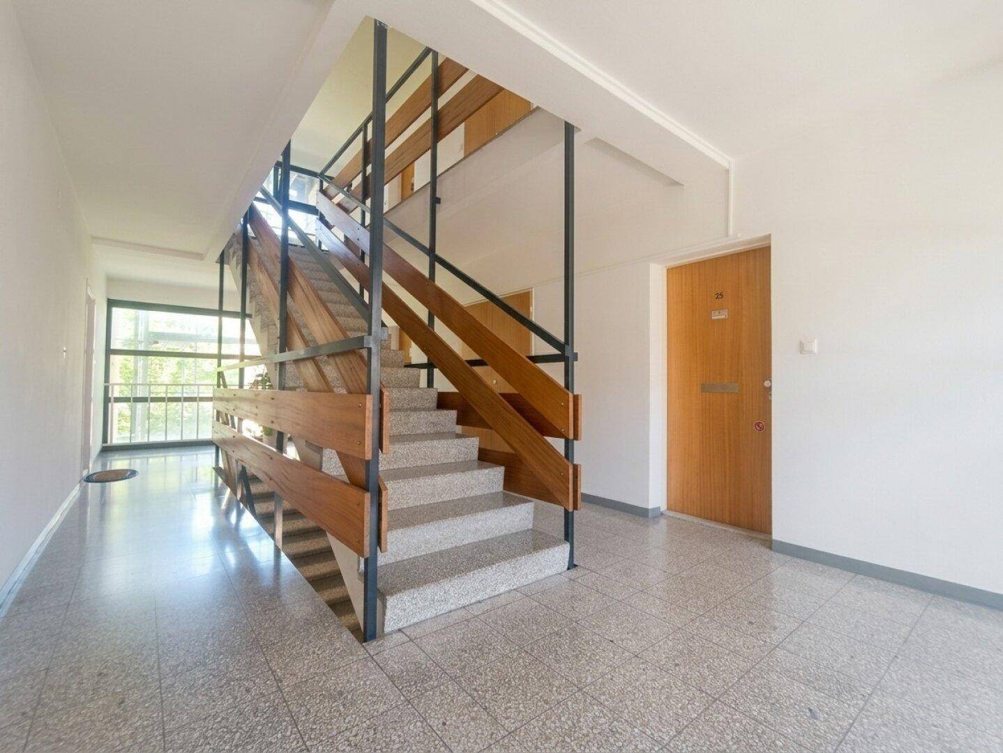 Treppenhaus mit Holzgeländer und Terrazzoboden, das die verschiedenen Etagen verbindet.