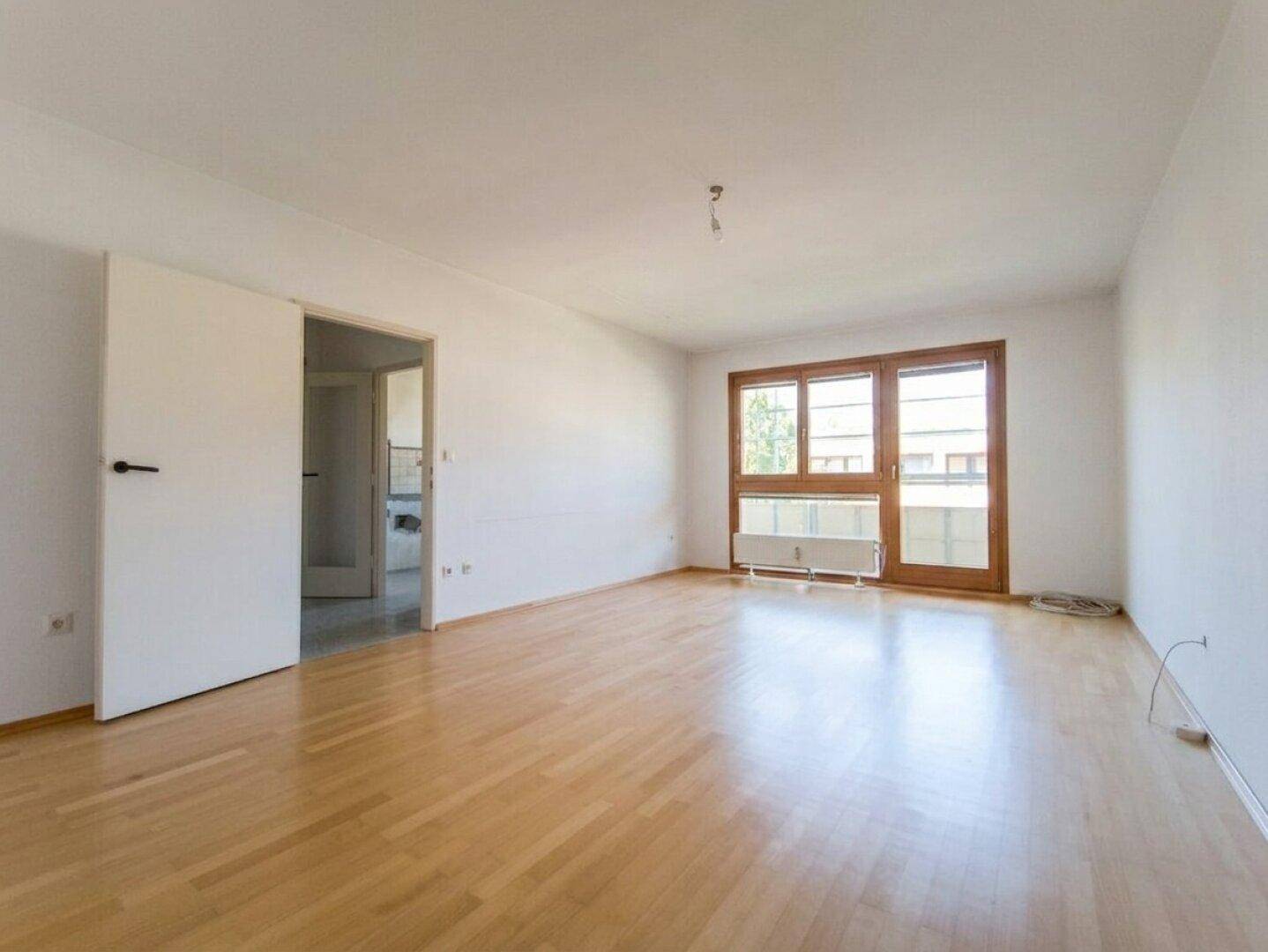 Helles, leeres Wohnzimmer mit hellem Parkettboden und Zugang zum Balkon.