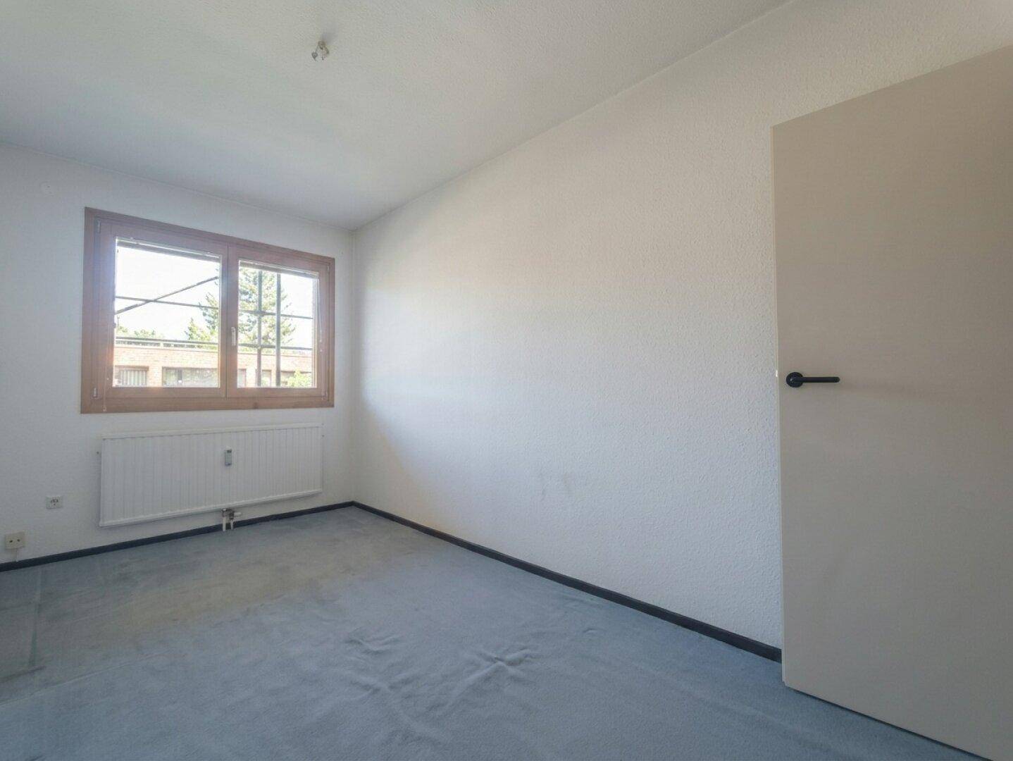 Leeres Schlafzimmer mit blauem Teppichboden und einem großen Fenster für viel Tageslicht.