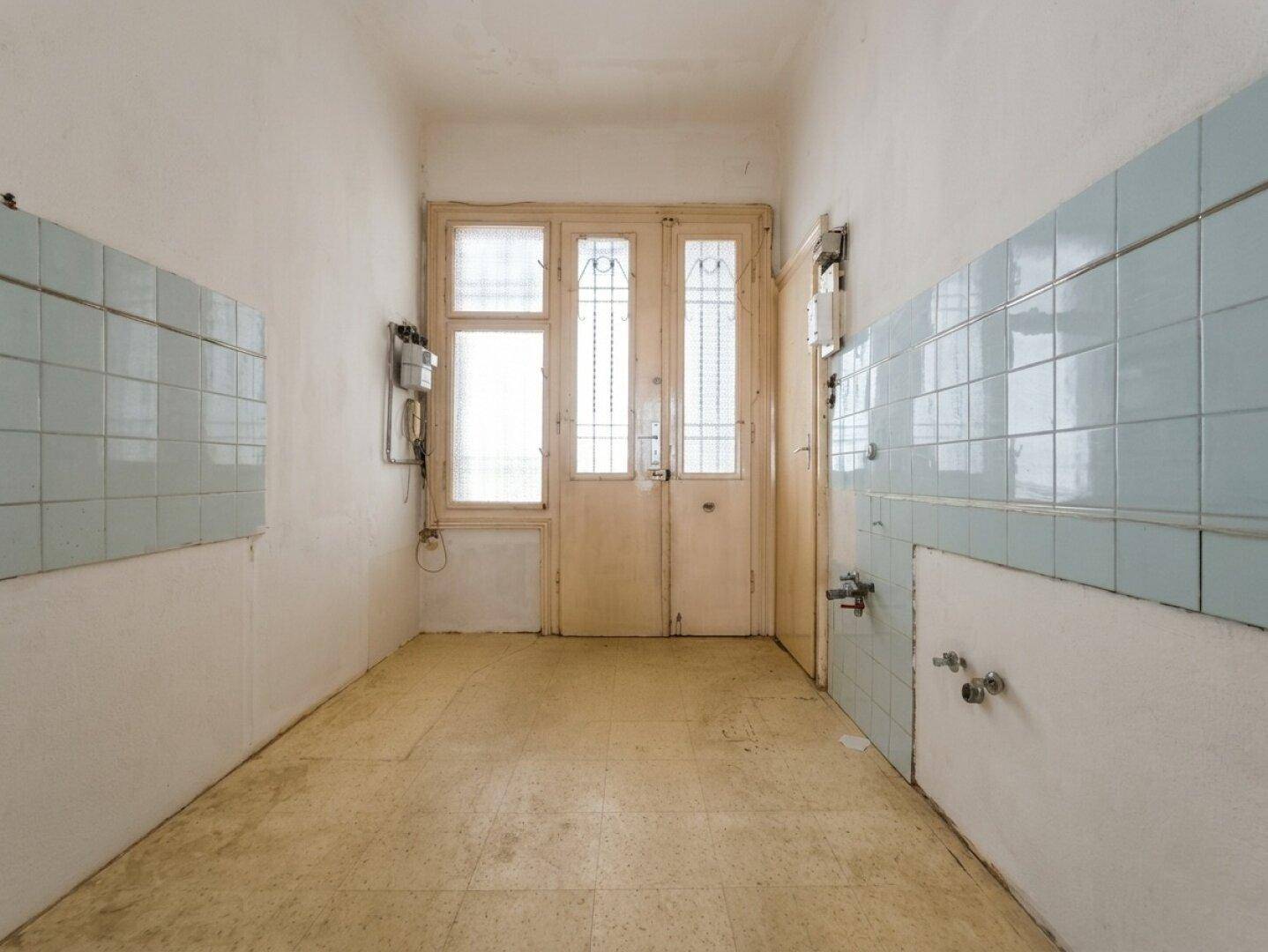 Unmöblierte Küche mit hellblauen Fliesen, Fenster und Tür, die auf eine Renovierung wartet.