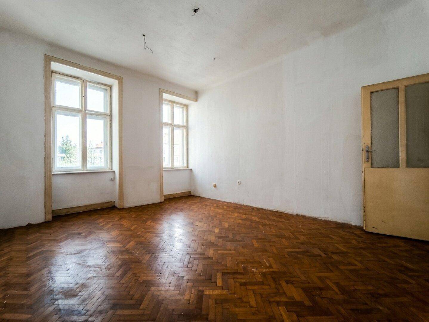 Leeres Zimmer mit Fischgrätparkettboden und zwei Fenstern, bereit für eine Renovierung.