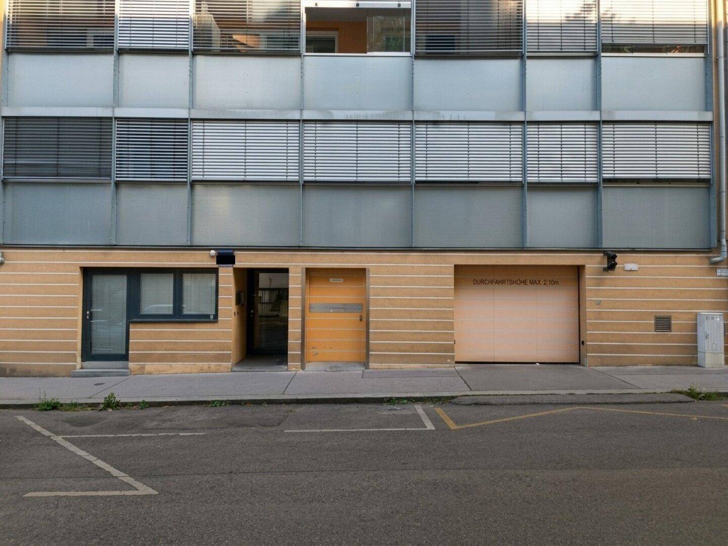 Fassade eines modernen Gebäudes mit Garagentor, Eingangstür und Jalousien an den Fenstern der oberen Etagen.