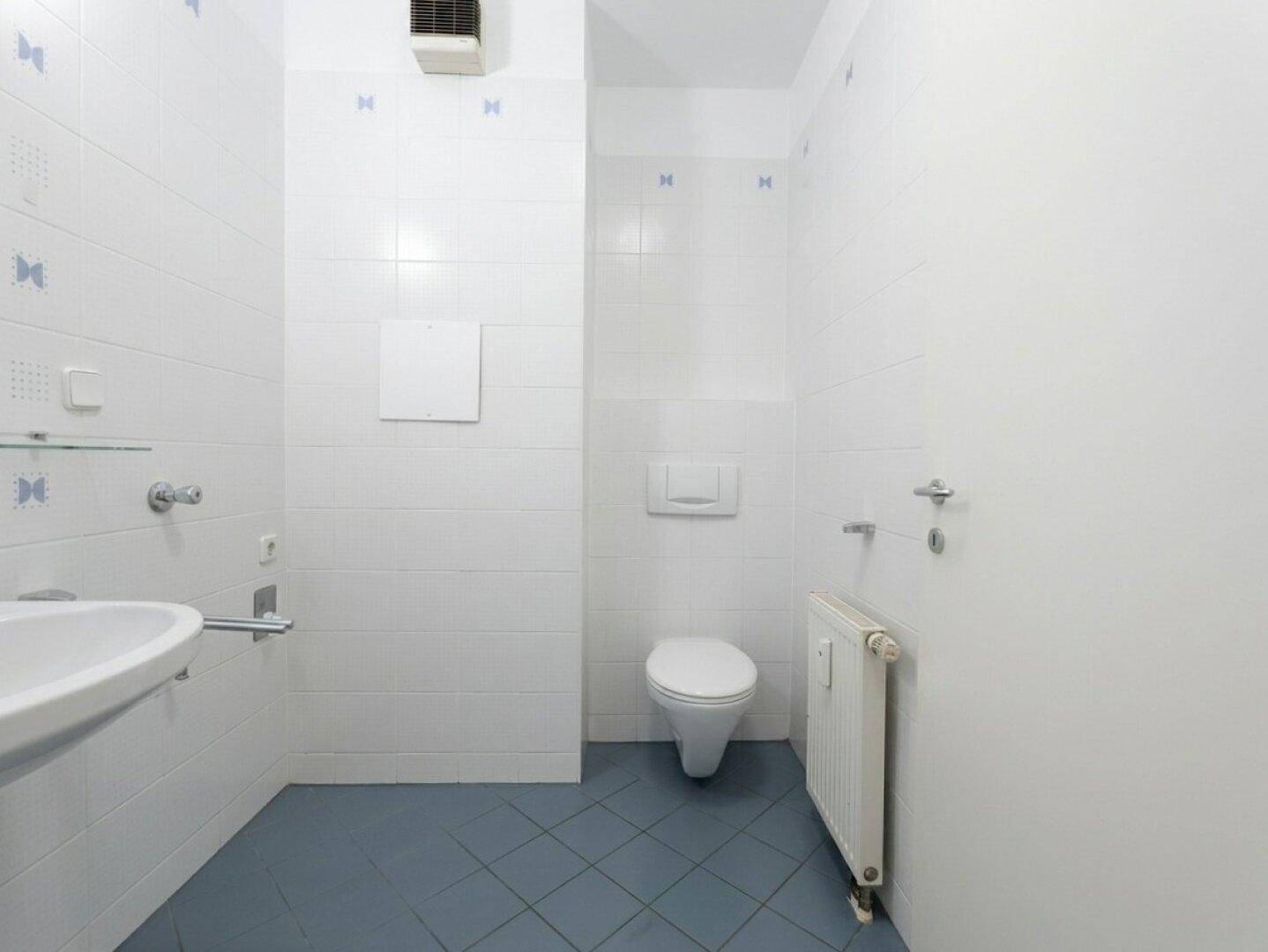 Separates WC mit weißen Fliesen, blauen Bodenfliesen, wandhängender Toilette und Waschbecken.