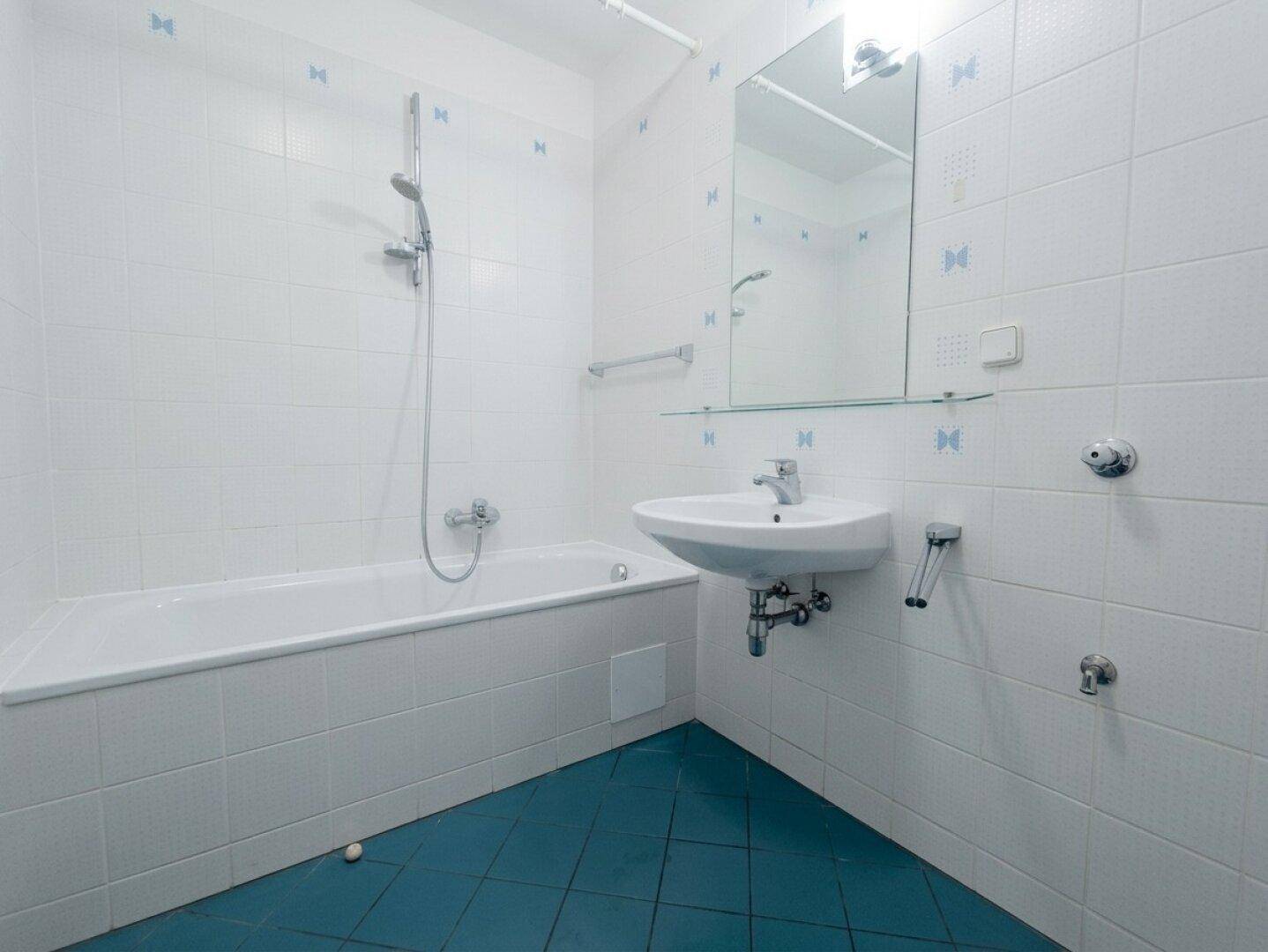 Badezimmer mit weißen Wandfliesen, blauen Bodenfliesen, Badewanne mit Duschfunktion und Waschbecken.