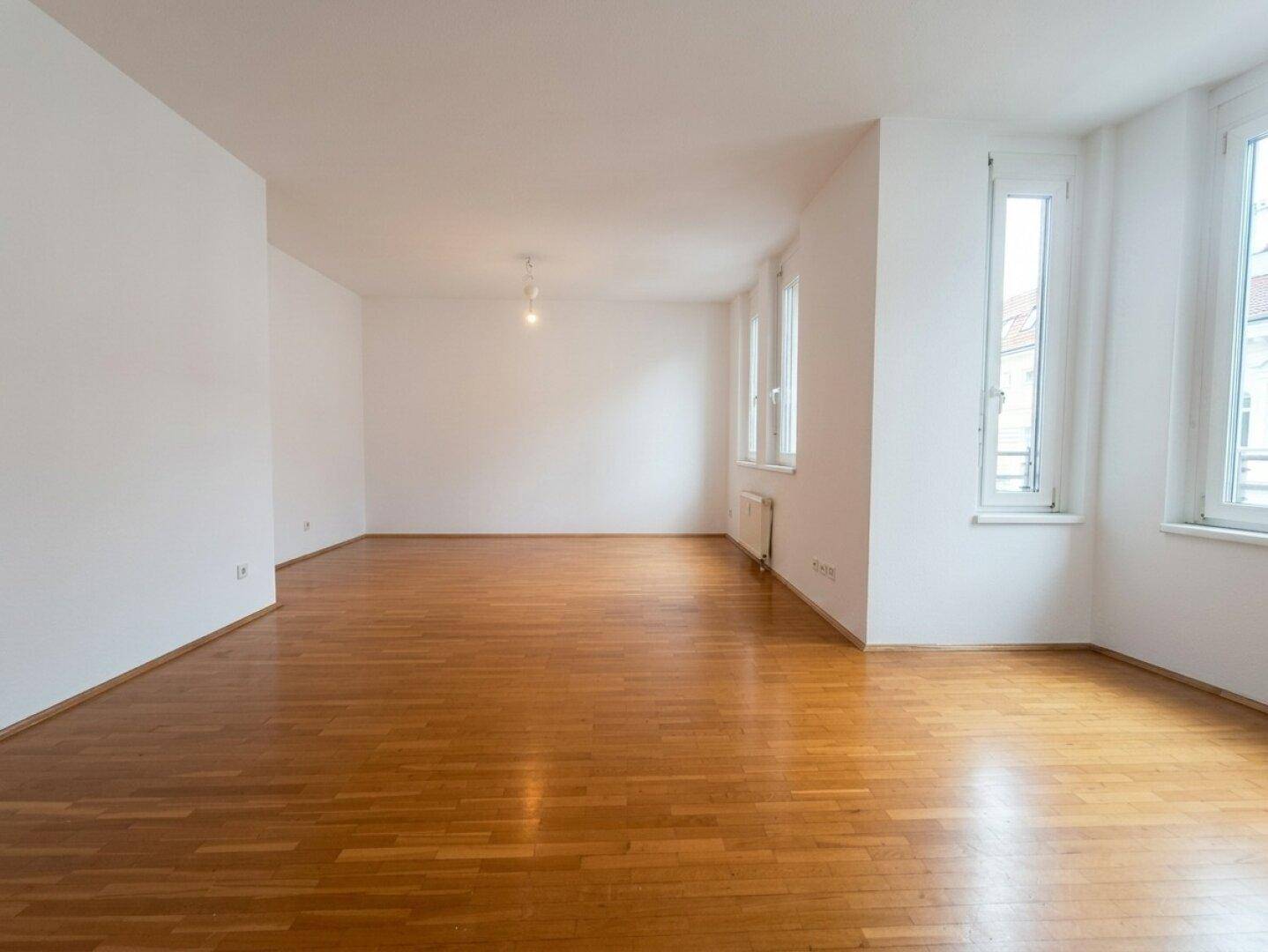 Geräumiges, leeres Wohnzimmer mit glänzendem Parkettboden und mehreren Fenstern für viel Tageslicht.