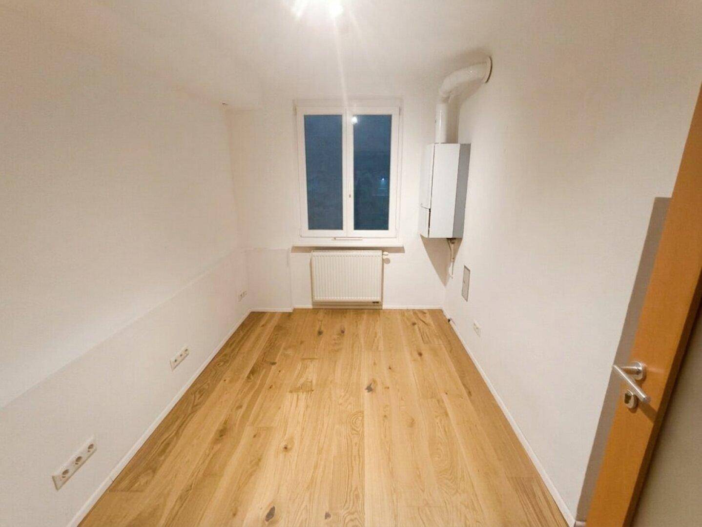 Kleiner Raum mit Fenster, Holzboden und wandmontiertem Heizkessel, ideal als Hauswirtschaftsraum.