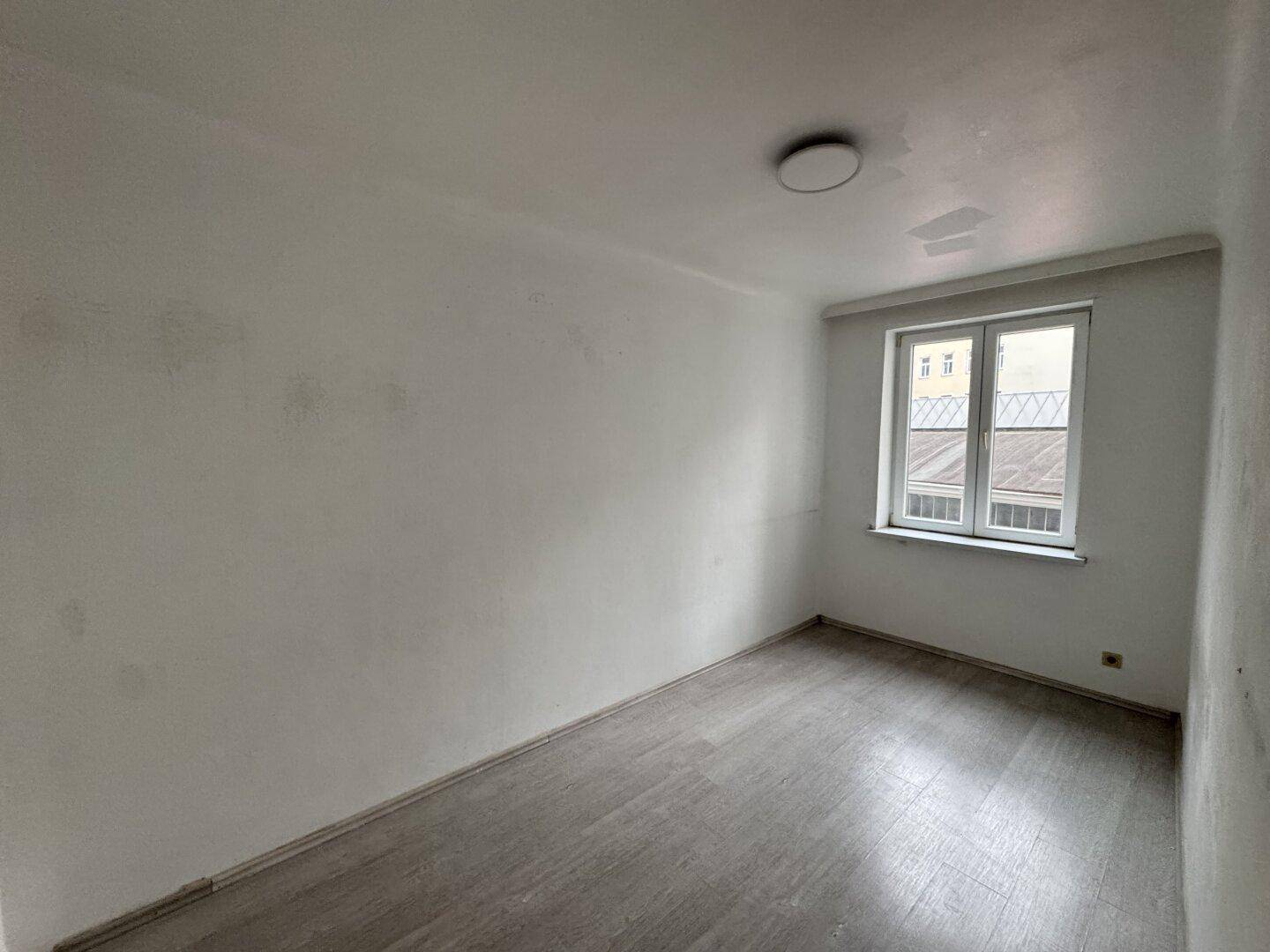 Leeres Zimmer mit hellgrauen Wänden, Laminatboden und einem Fenster, das einen Blick auf die Stadt bietet.