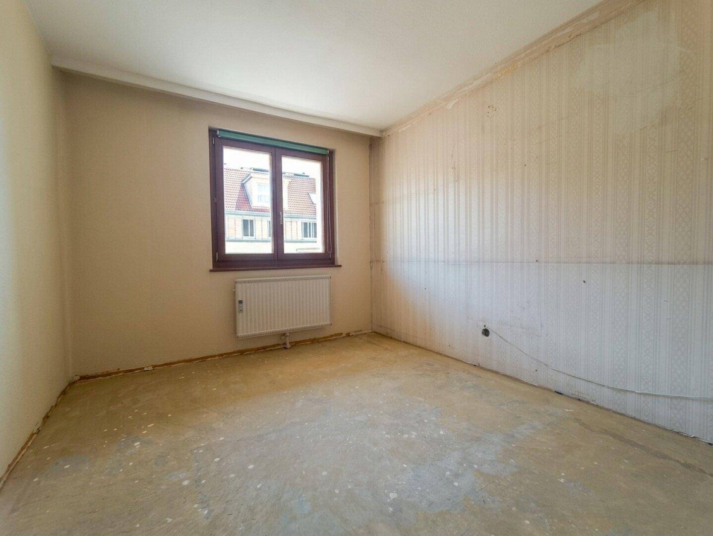 Leeres Schlafzimmer mit abgenutzter Tapete und Fenster, das auf eine Renovierung wartet.