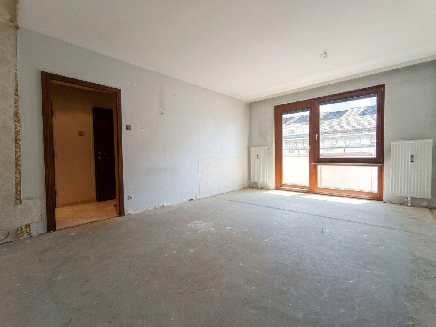 Unmöblierter Raum mit Betonboden und großen Fenstern, bereit für die Renovierung.