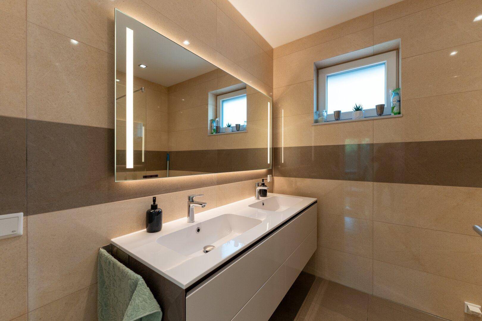 Elegantes Badezimmer mit Doppelwaschbecken, beleuchtetem Spiegel und beigefarbenen Fliesen.