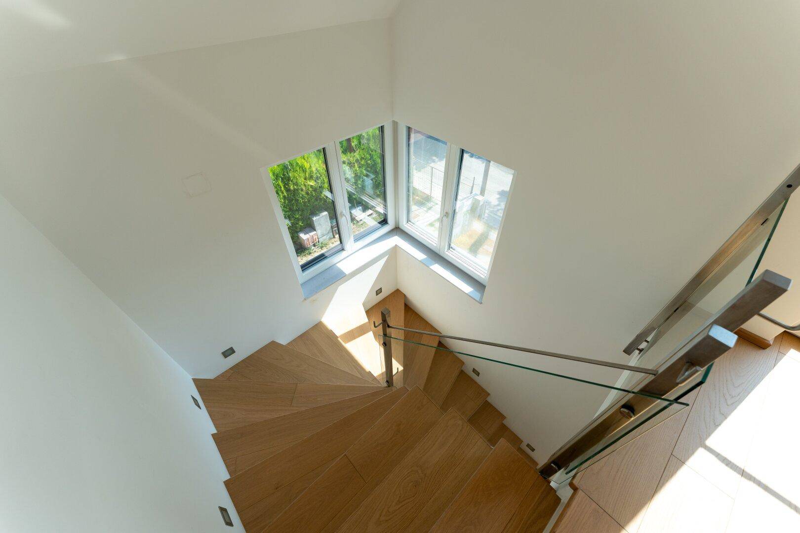 Moderne Holztreppe mit Glasgeländer und großen Fenstern, die viel Licht hereinlassen.