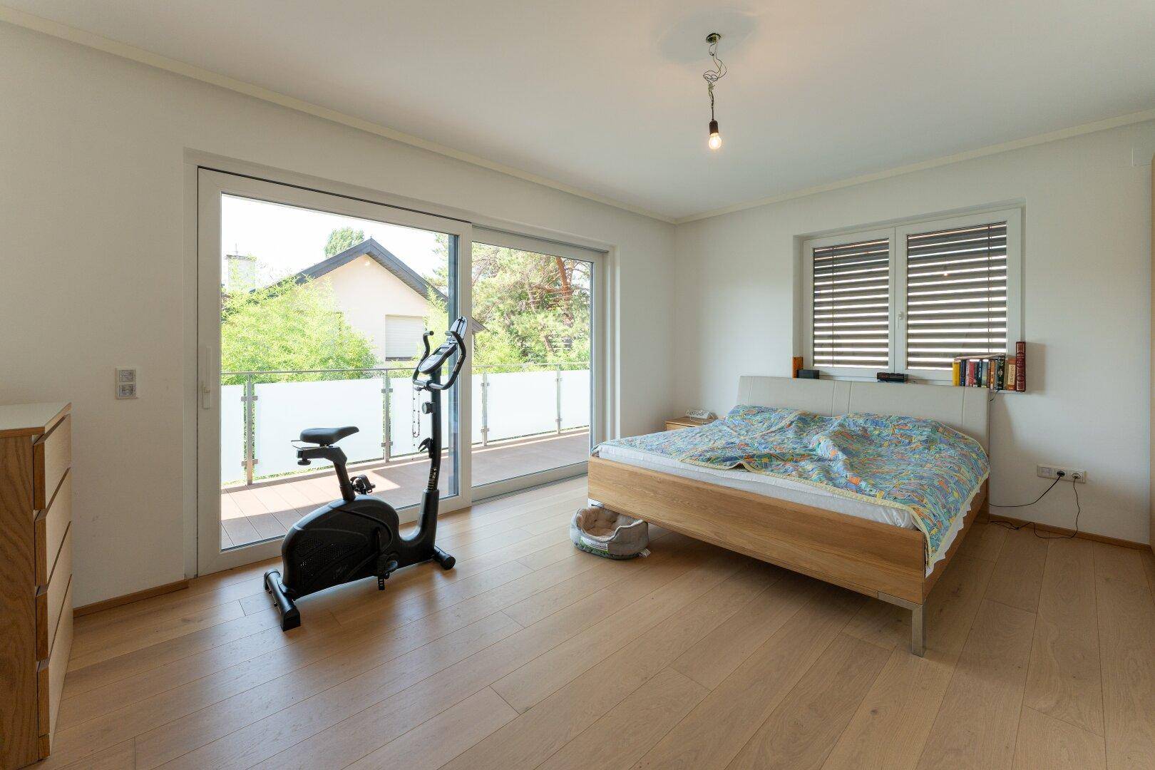 Geräumiges Schlafzimmer mit Holzboden, Zugang zum Balkon und einem Heimtrainer.