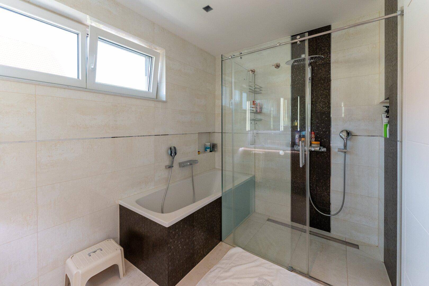 Modernes Badezimmer mit begehbarer Dusche, Badewanne und Fenster, hell gefliest.