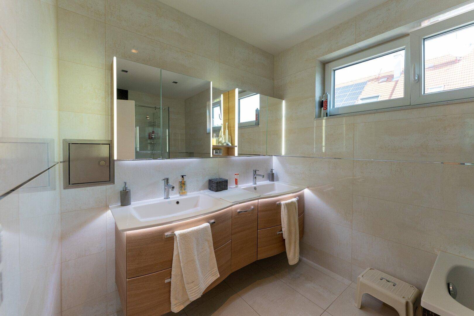 Helles Badezimmer mit Doppelwaschbecken, beleuchtetem Spiegelschrank und Fenster.